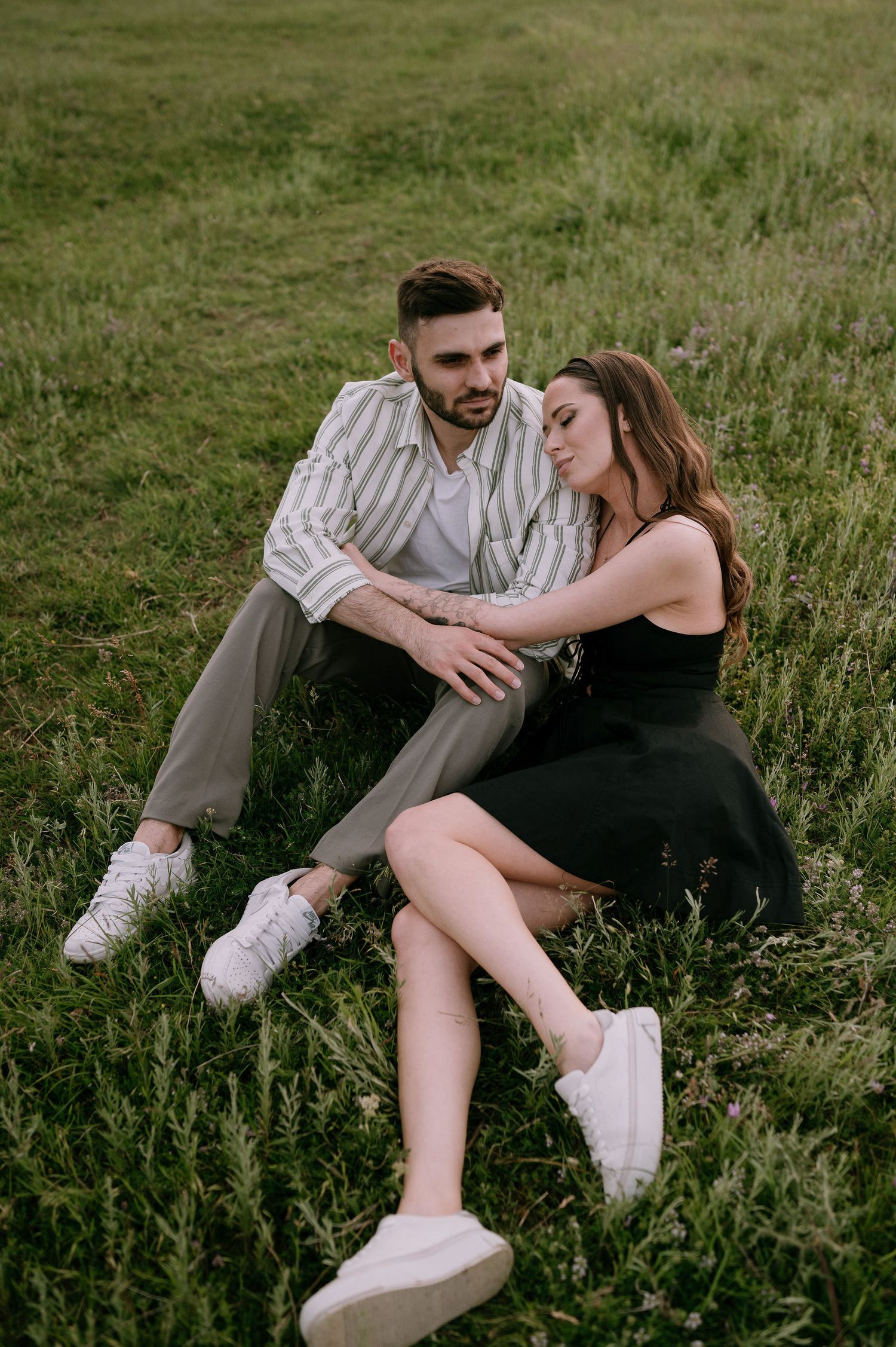 A couple relaxes together on grassy hillside during romantic outdoor photoshoot at sunset.
