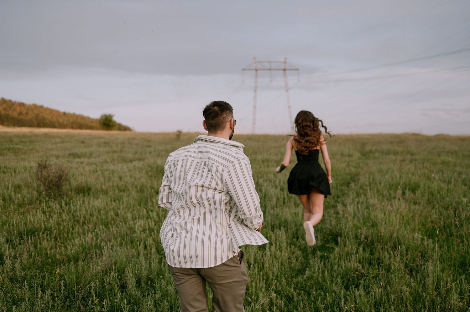 Two people walk through a green meadow with electrical transmission towers visible in the distance on an overcast day.
