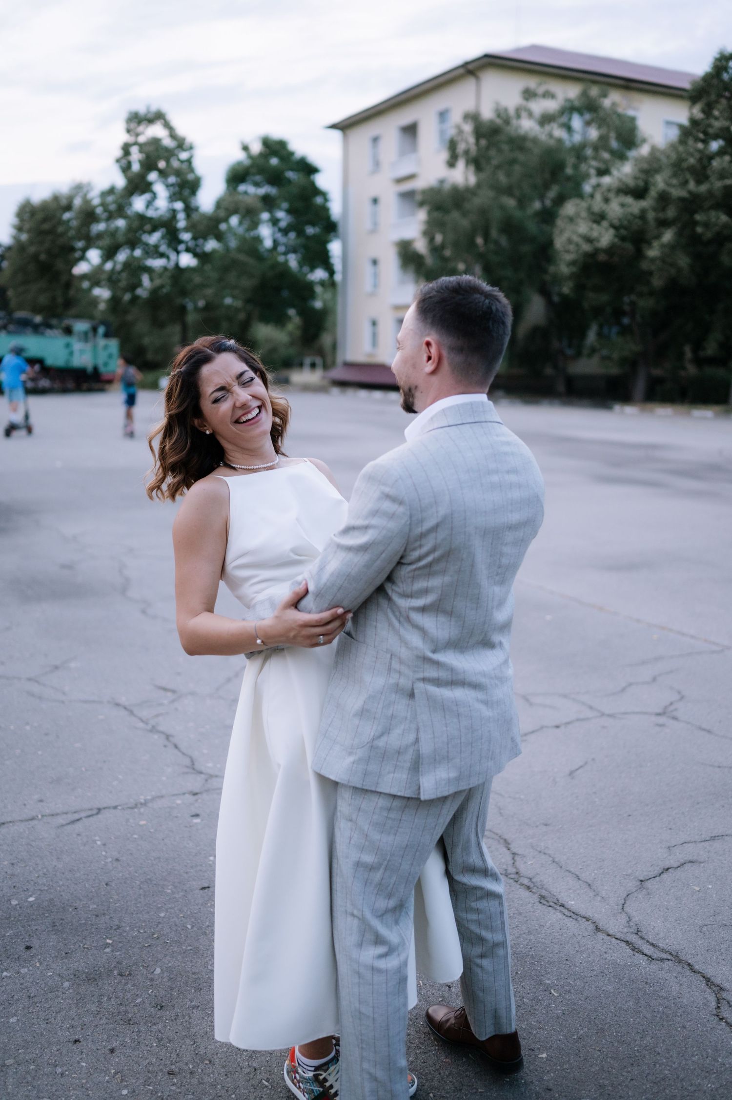 A couple dances playfully in a parking lot, the woman wearing a white dress and man in a gray suit.