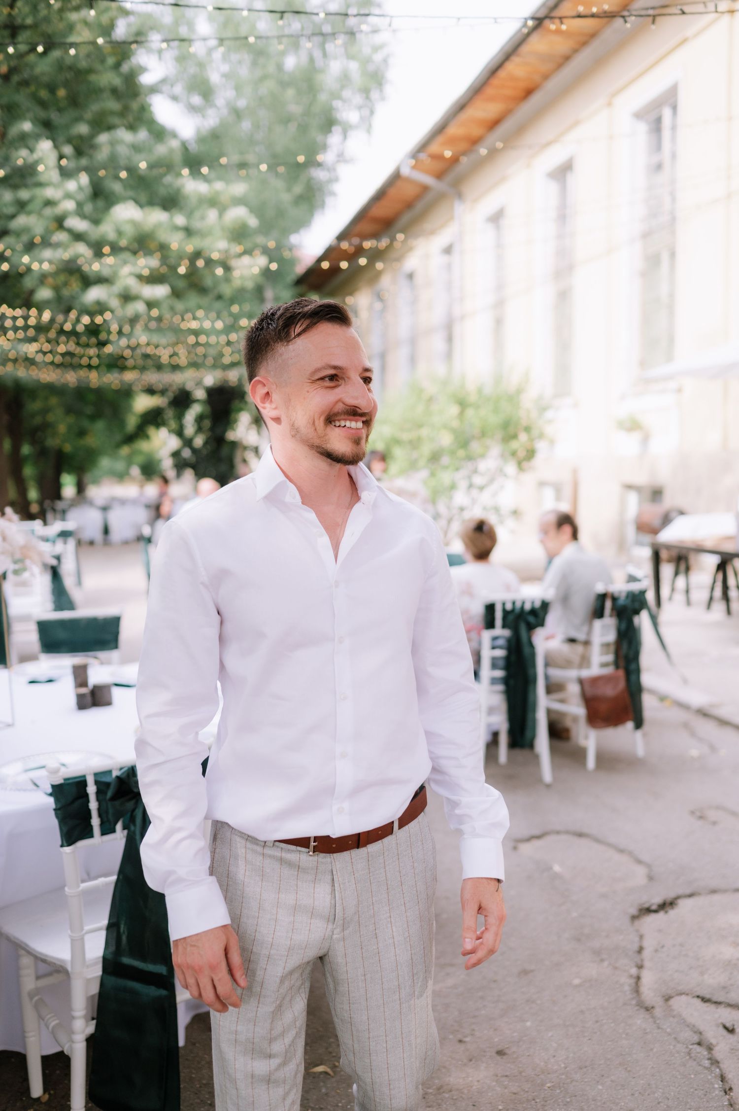 A person wearing a white dress shirt and light gray vest stands at an outdoor wedding reception venue with string lights.