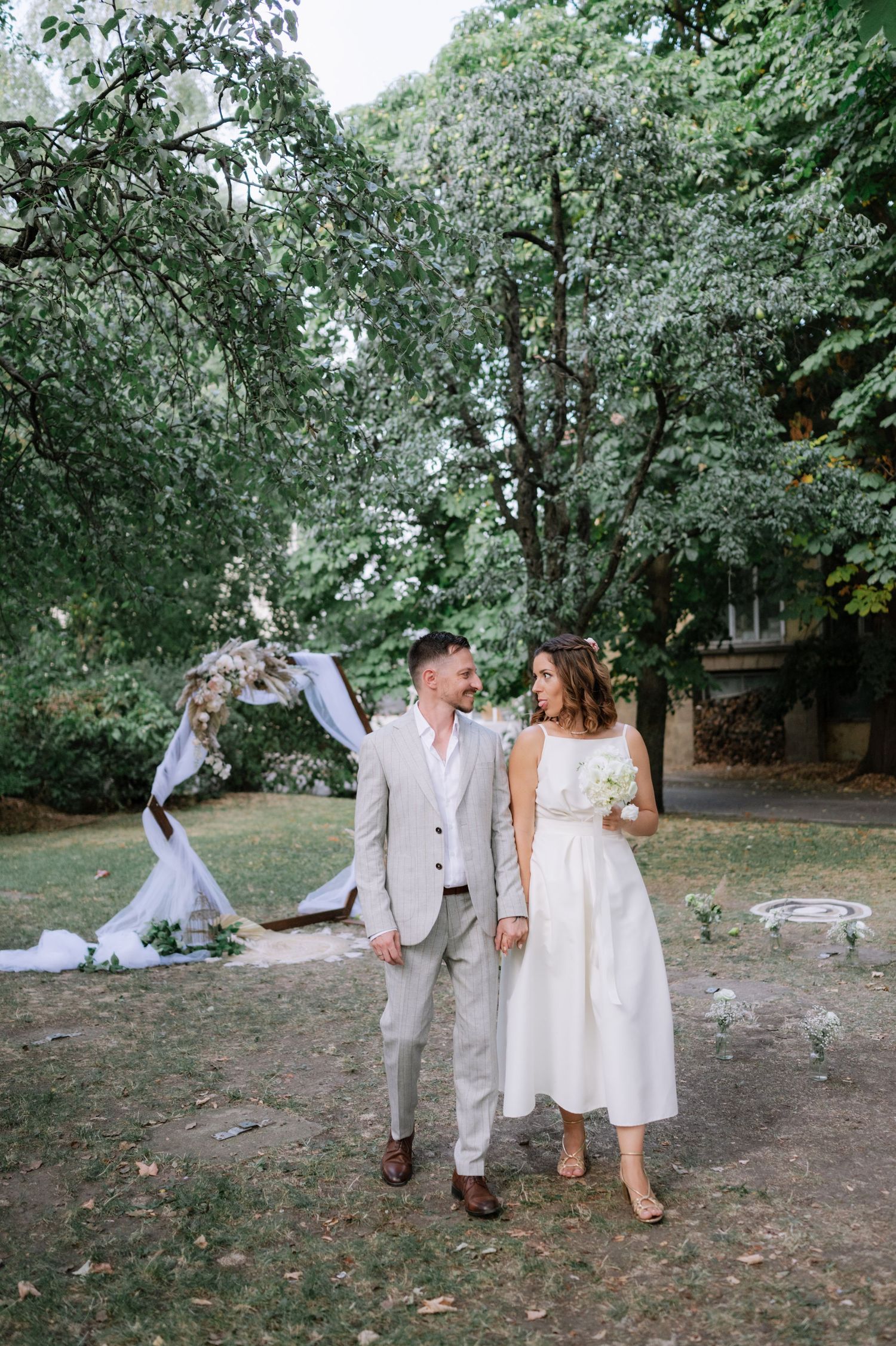 A couple walks hand in hand through a lush green park setting during what appears to be a casual outdoor celebration.