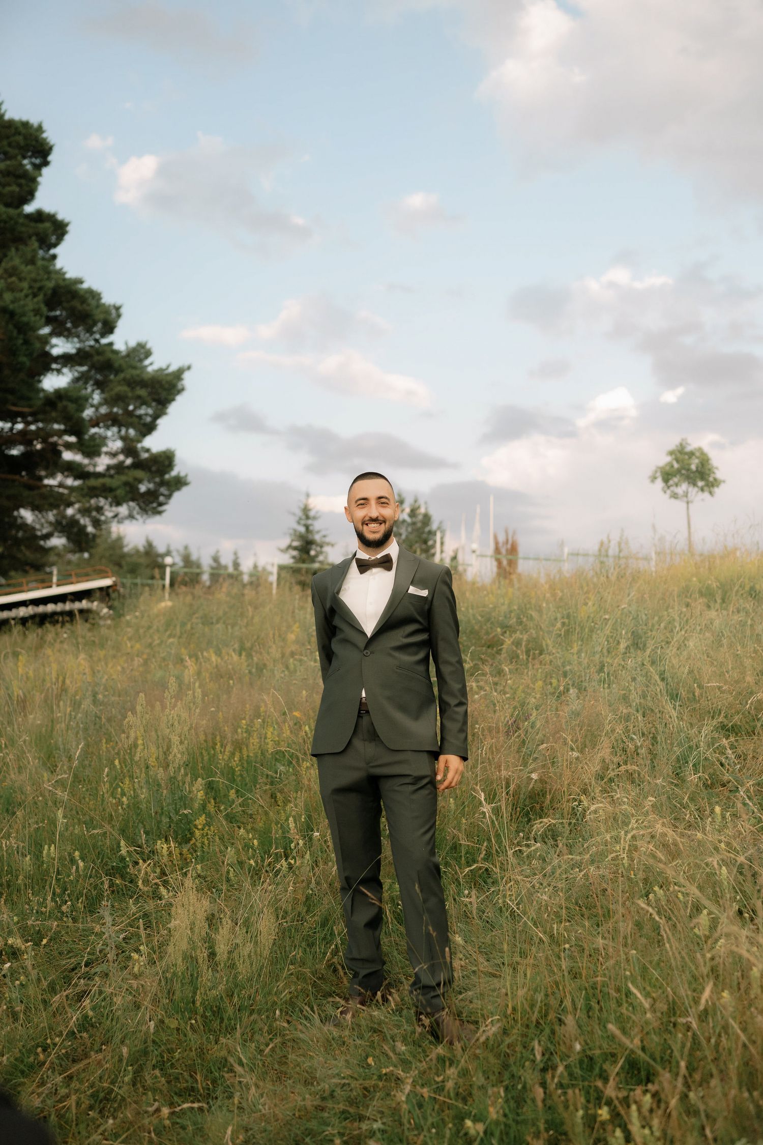 A figure in a dark suit stands in a scenic meadow with tall grass and trees under a cloudy sky.