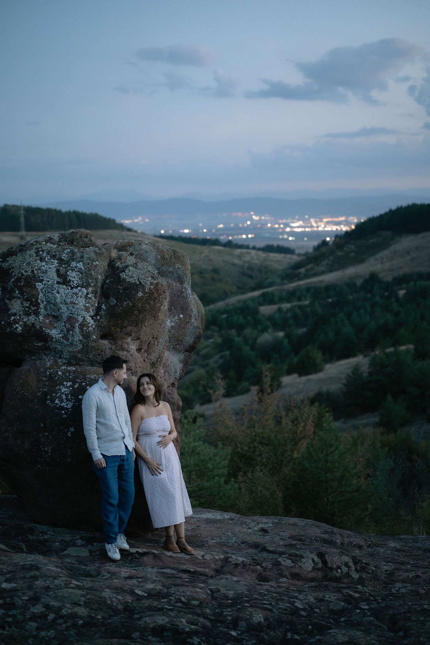 A couple sits together on a rocky overlook at dusk with a scenic mountain valley and city lights in the distance.