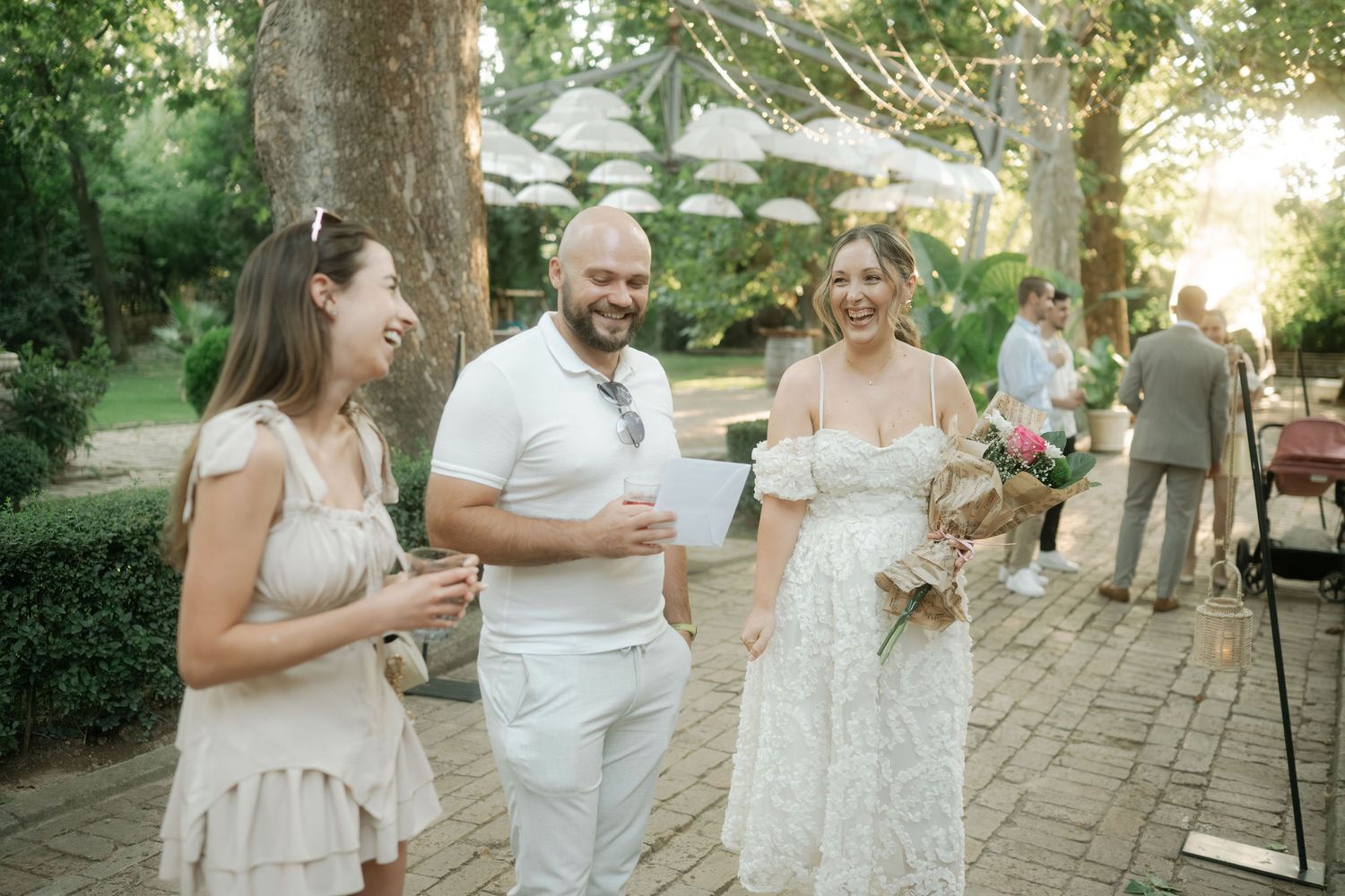 Wedding party gathered outdoors under trees on a brick patio during a sunny celebration.