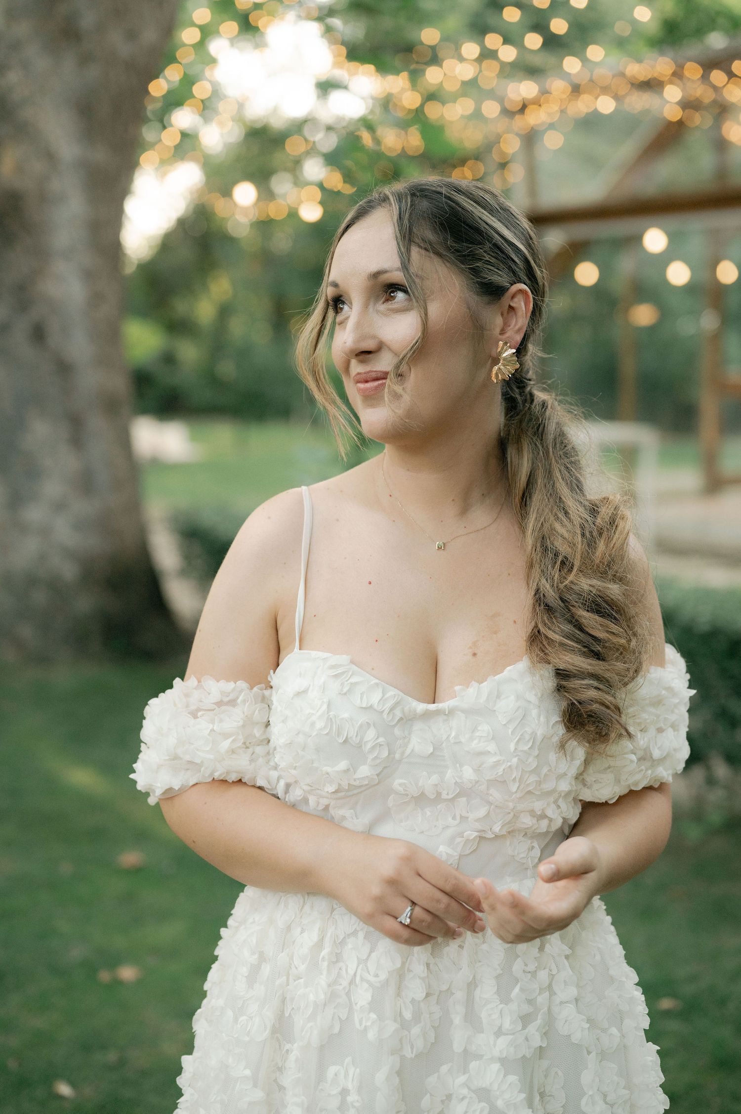 A person in a delicate off-shoulder white lace wedding dress poses outdoors with soft bokeh lighting in the background.