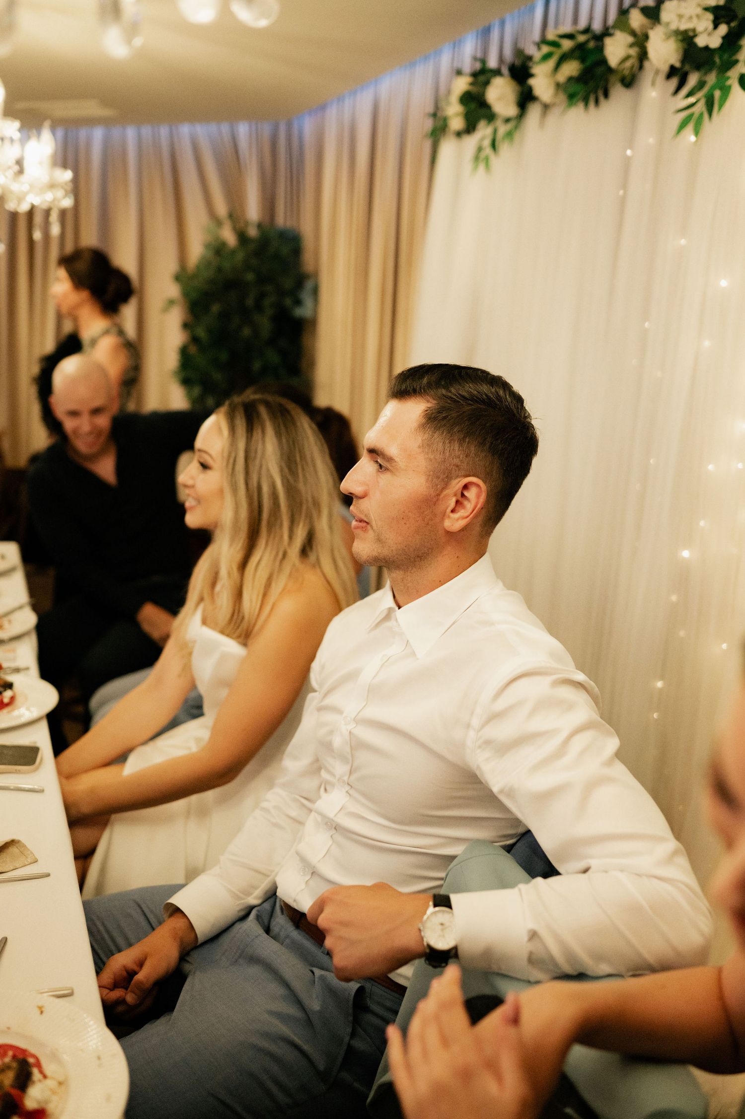 Wedding reception scene with guests seated at decorated tables with floral arrangements and twinkle lights.