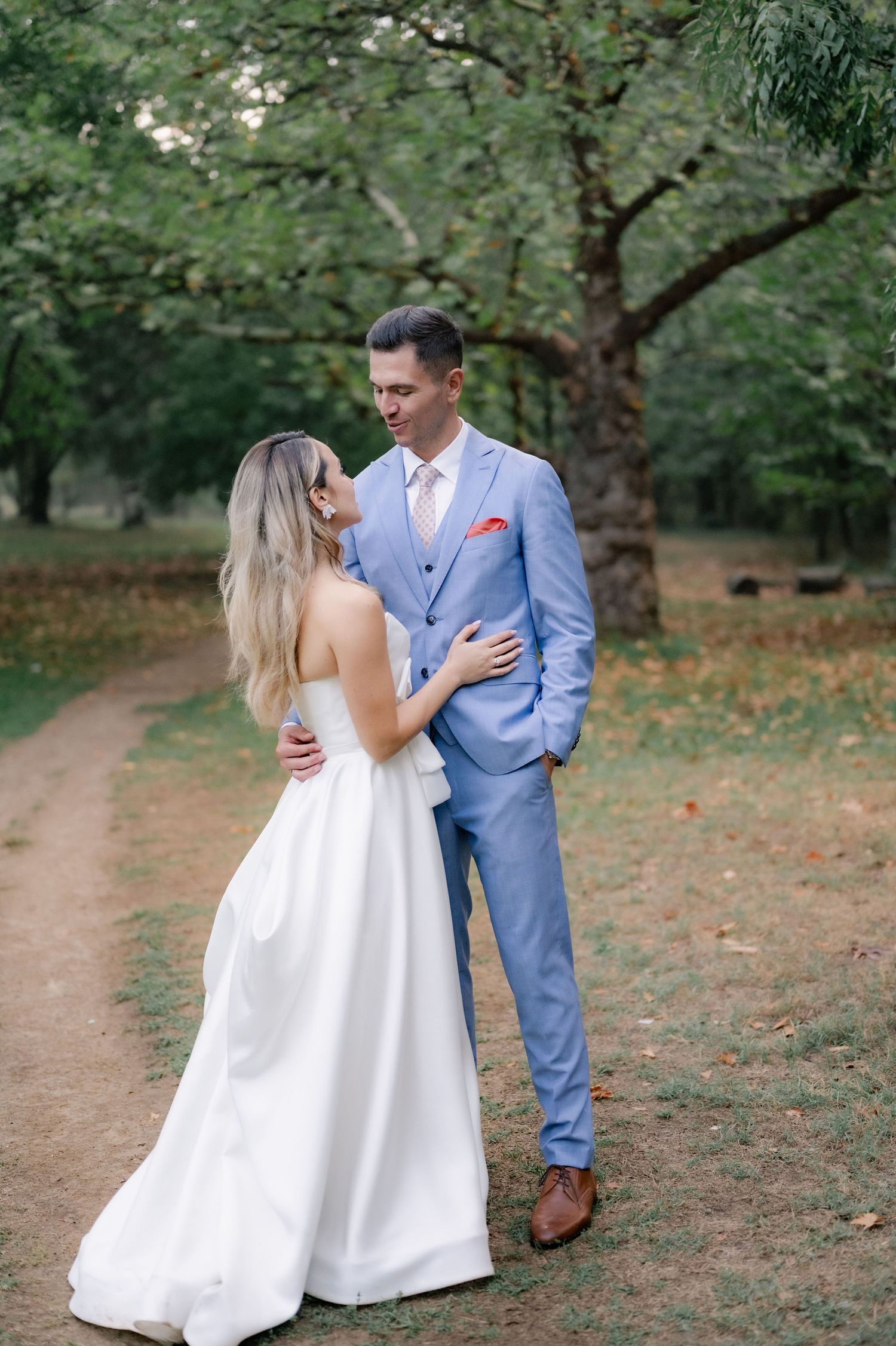 A couple in a blue suit and white dress share intimate moments during an outdoor photo session in a garden setting.