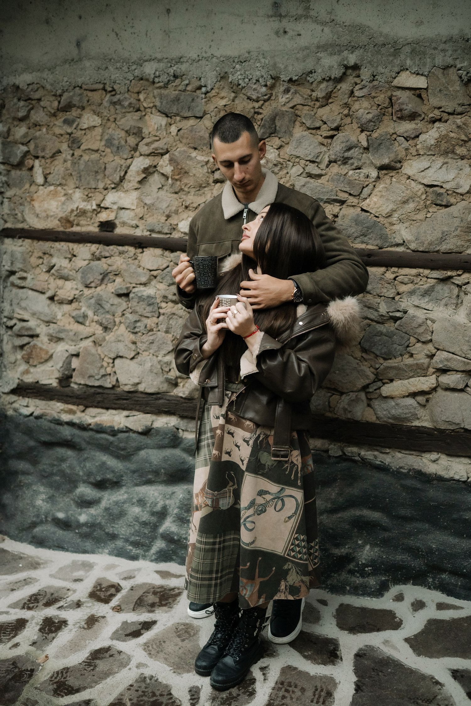 A person in traditional Scottish attire with a kilt and jacket stands against a rustic stone wall.