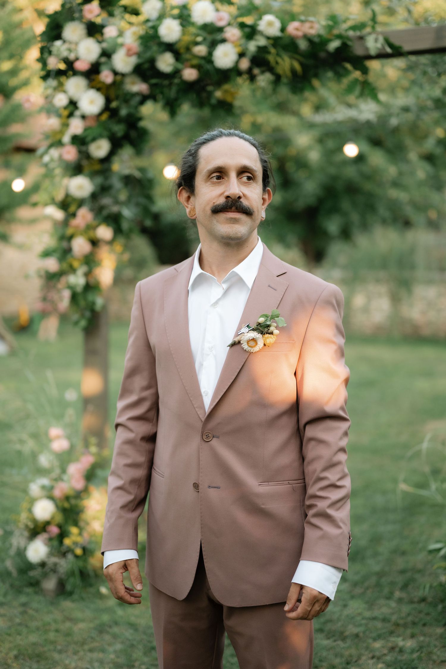 A person in a brown suit and white shirt poses in a garden setting with blooming roses in the background.