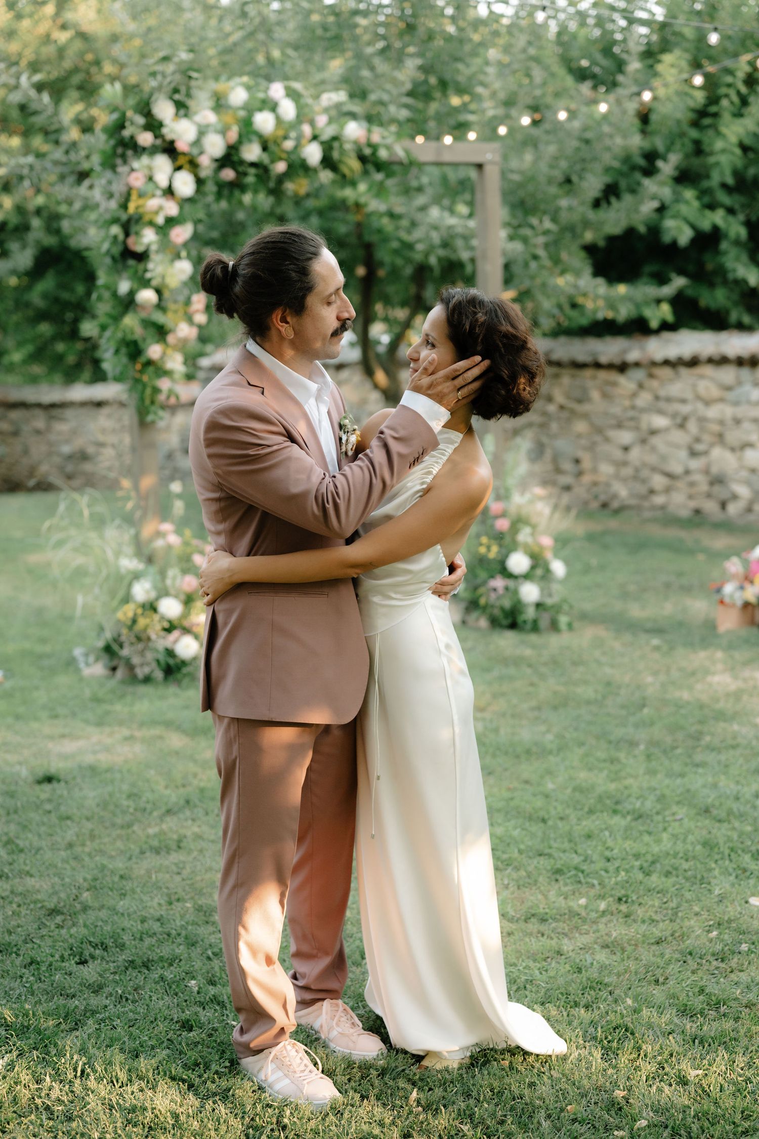 A couple shares intimate moments in a garden setting while wearing brown and cream colored wedding attire.