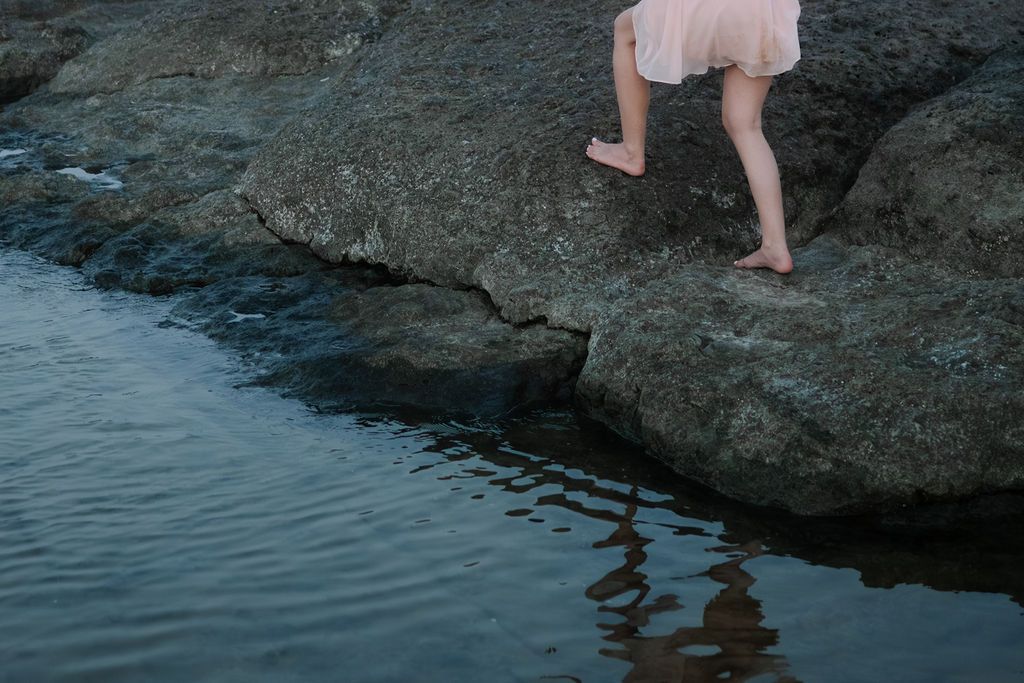 A person stands on rocky cliffs overlooking the ocean while taking photos at sunset.