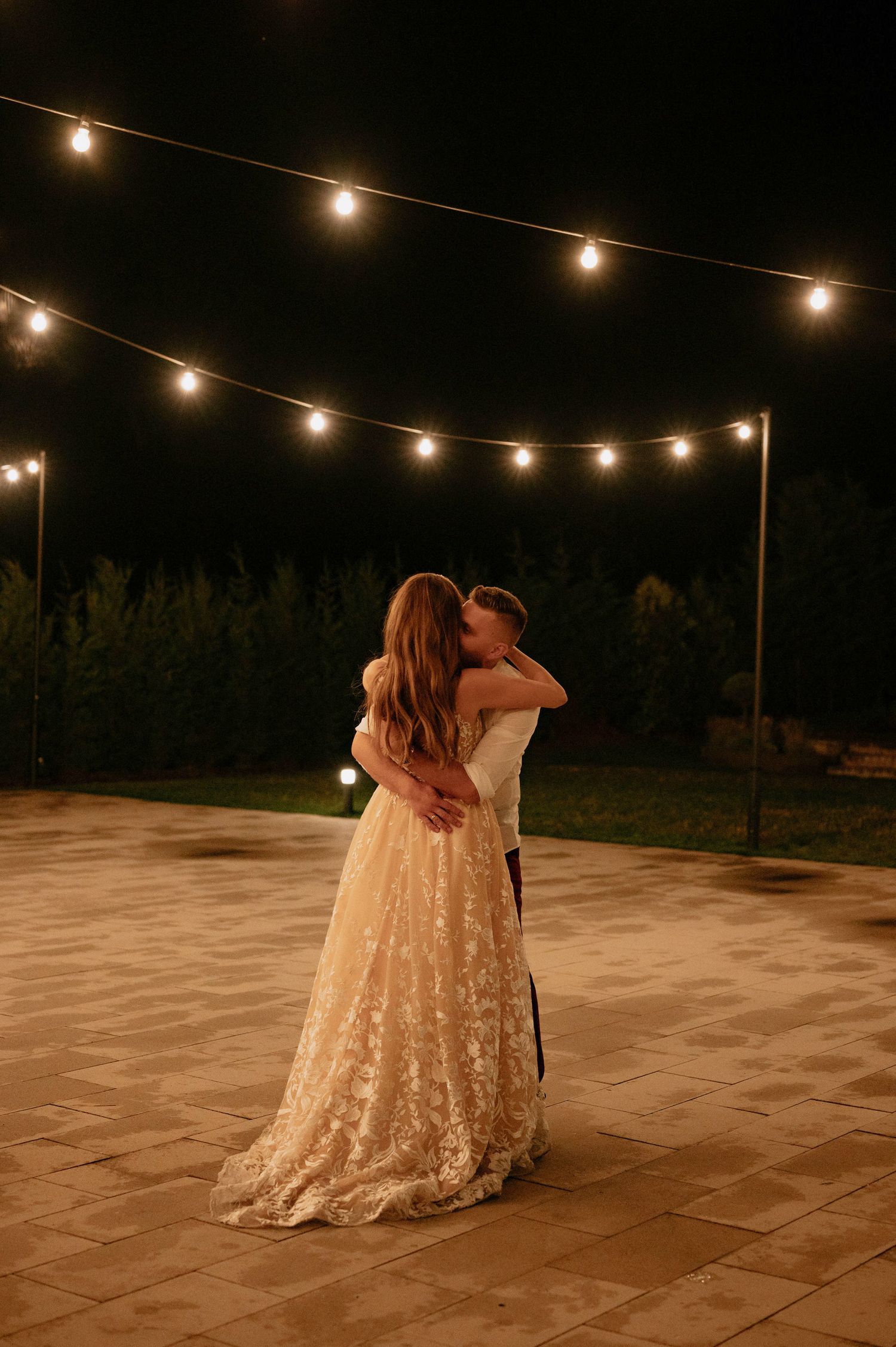 A romantic dance under string lights on a stone patio at night with a flowing champagne-colored lace dress.