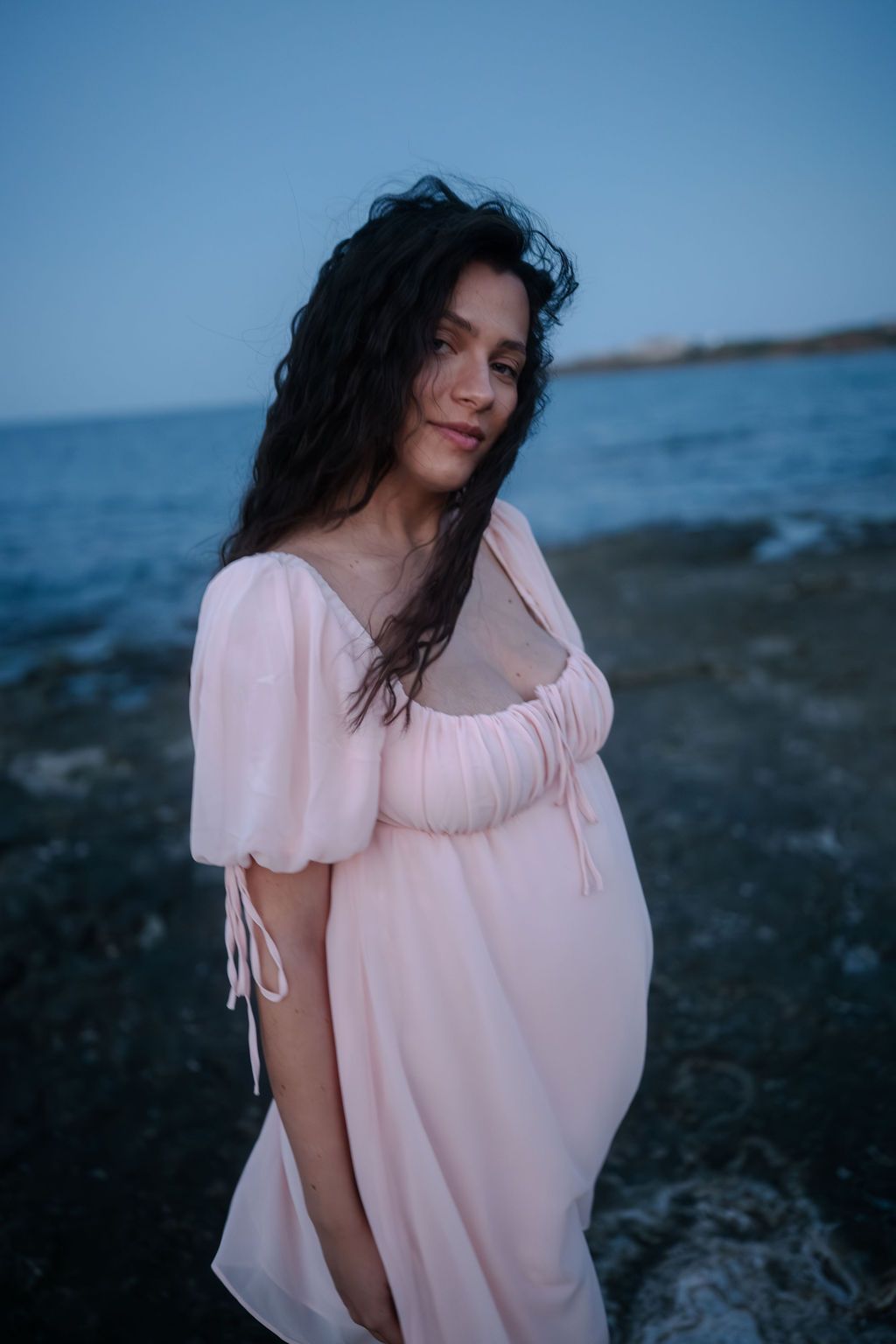 Dreamy coastal portrait series in pink dress against moody blue seaside backdrop at dusk.