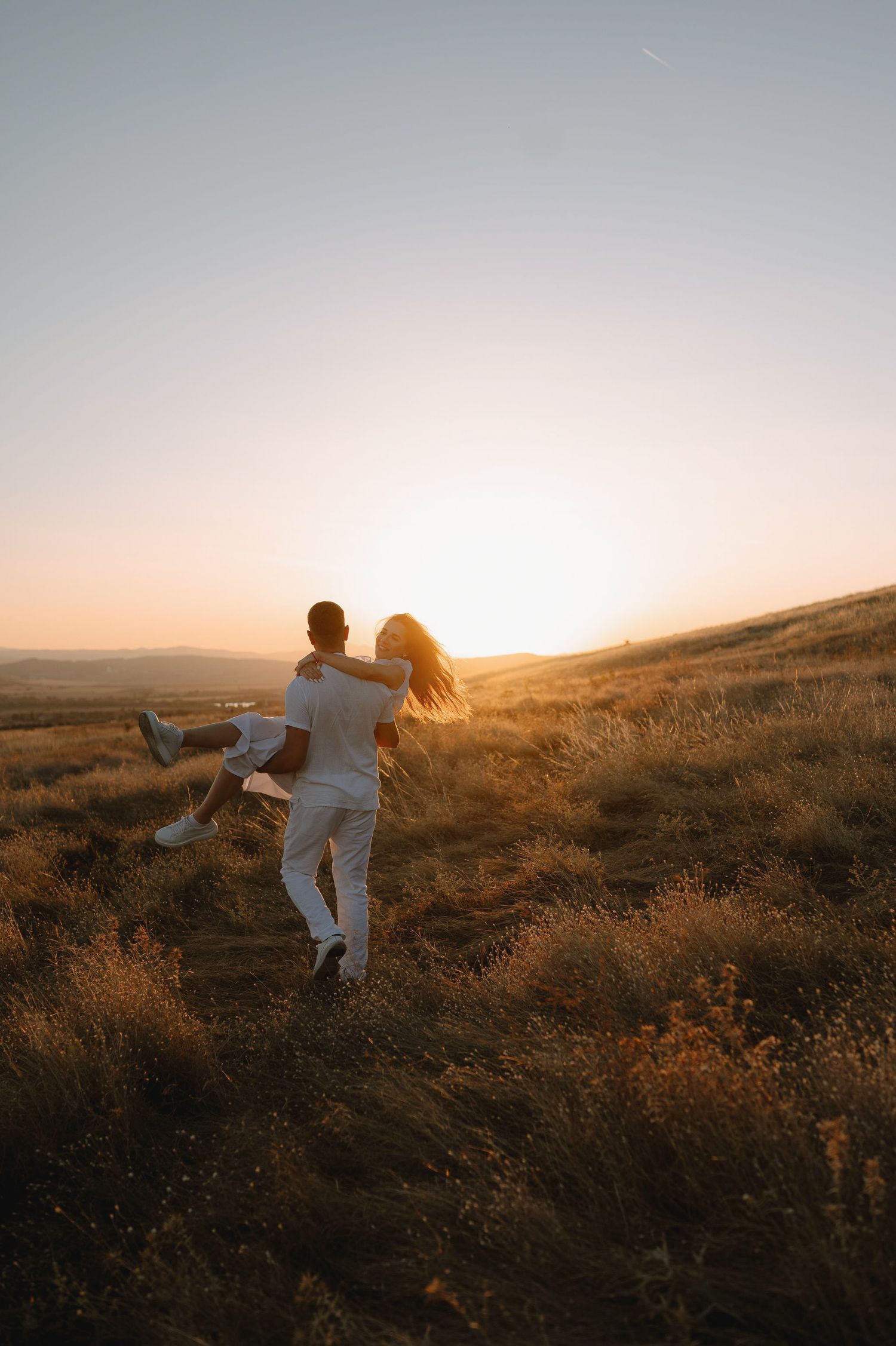 A couple runs through a golden field at sunset, their silhouettes backlit against the warm glowing sky.