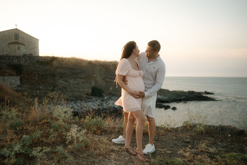 A romantic coastal photoshoot at sunset shows a couple embracing on rocky cliffs overlooking the ocean.