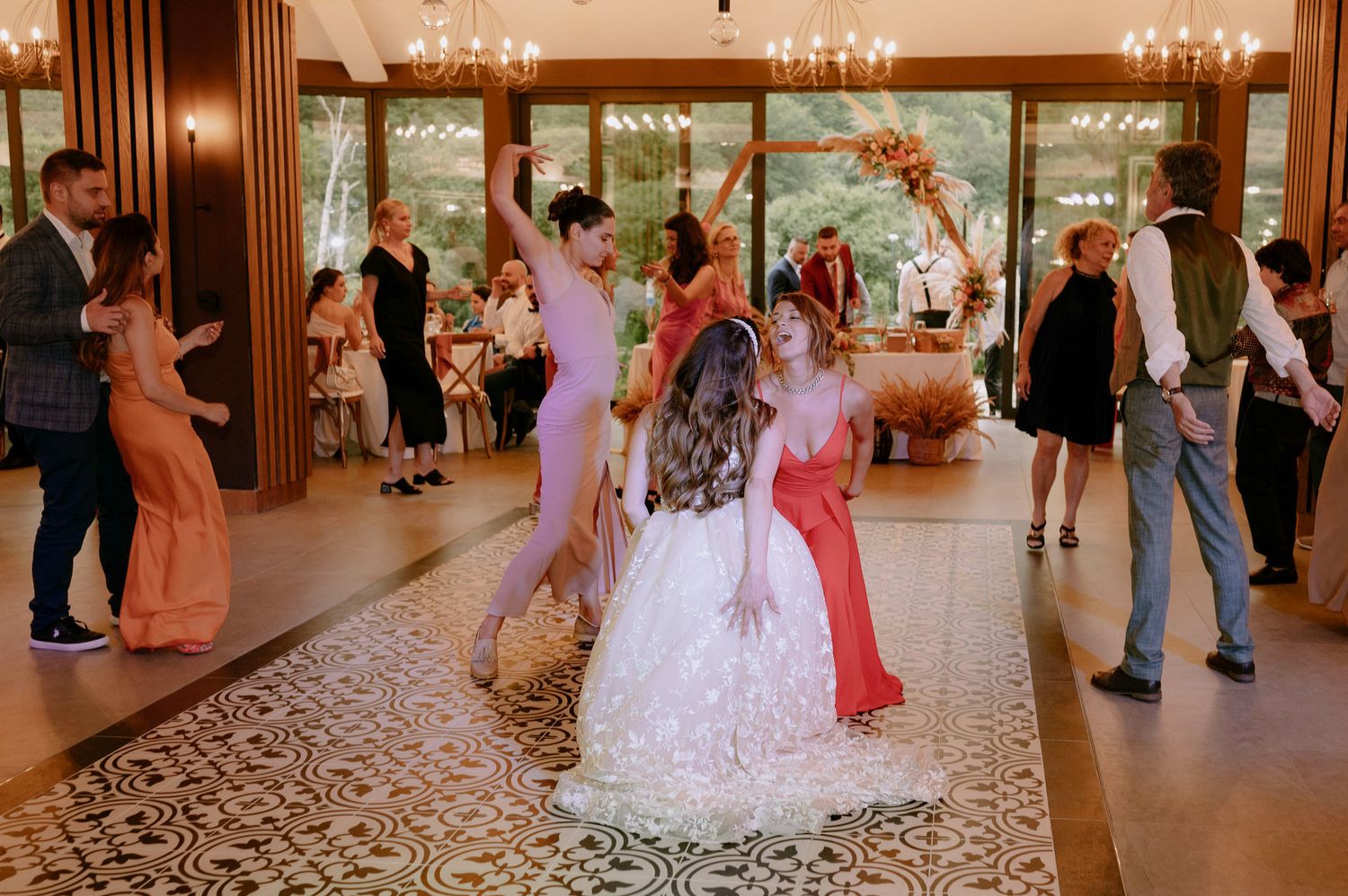 Wedding guests dance and celebrate on an elegant ballroom floor with chandeliers and large windows.