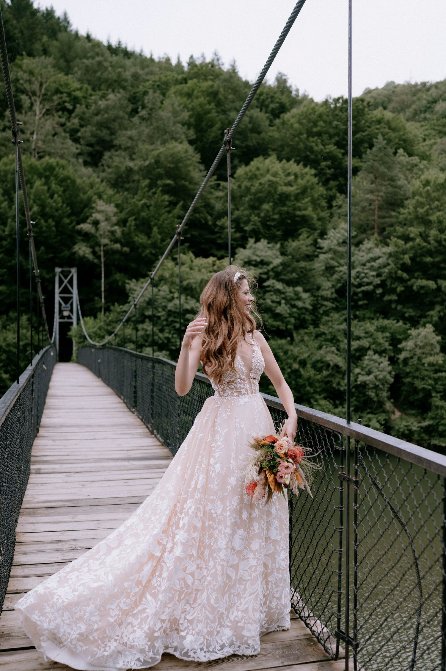 A bride in a lace wedding dress walks across a rustic wooden suspension bridge surrounded by lush forest.