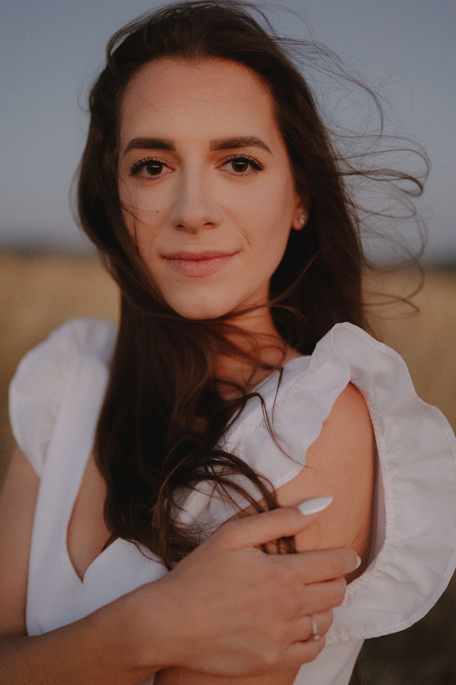 Portrait of a woman with long dark hair in a white top smiling at sunset with wind blowing through her hair.