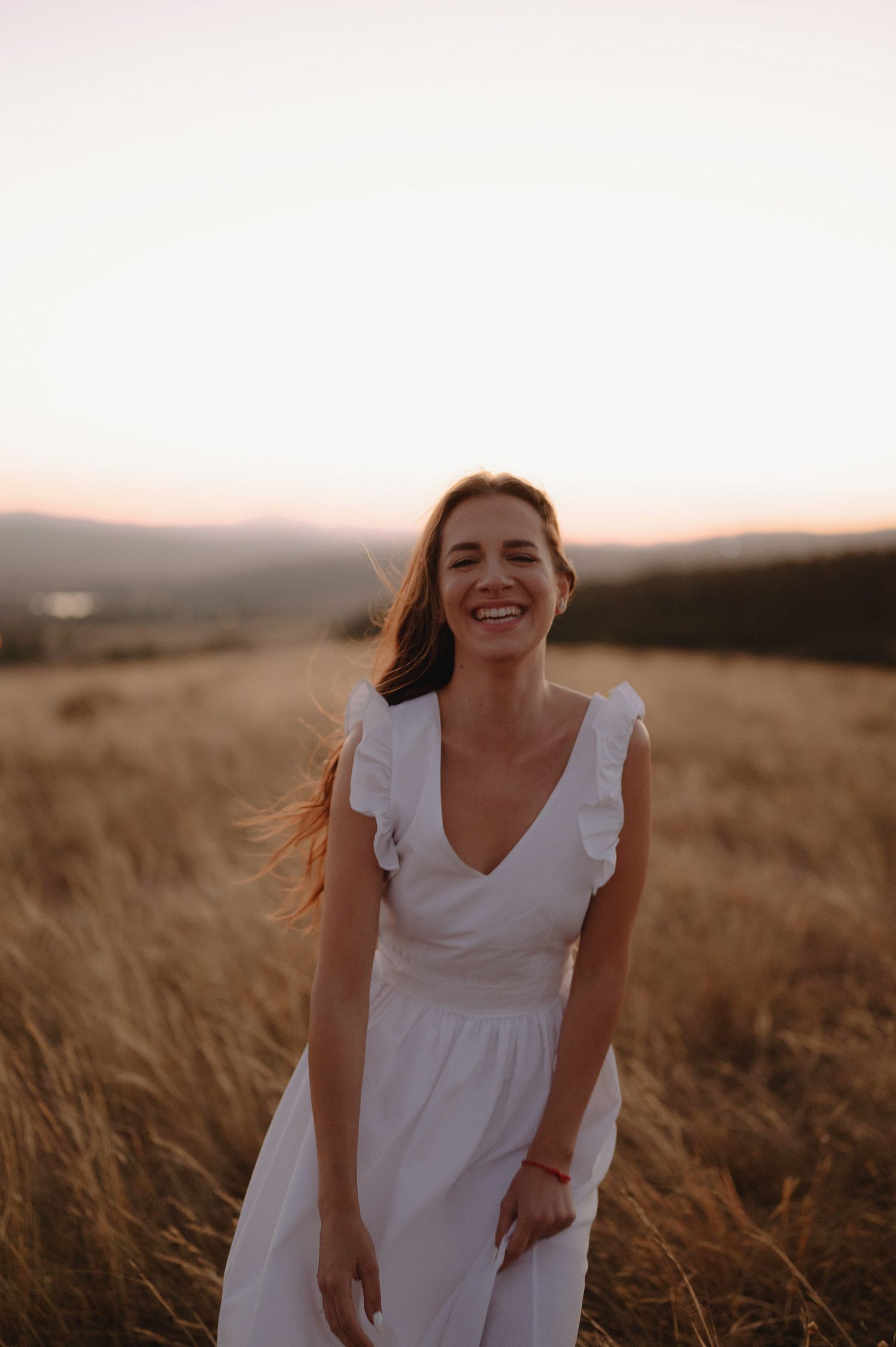 A person in a white dress stands in a golden wheat field at sunset, smiling and leaning forward.