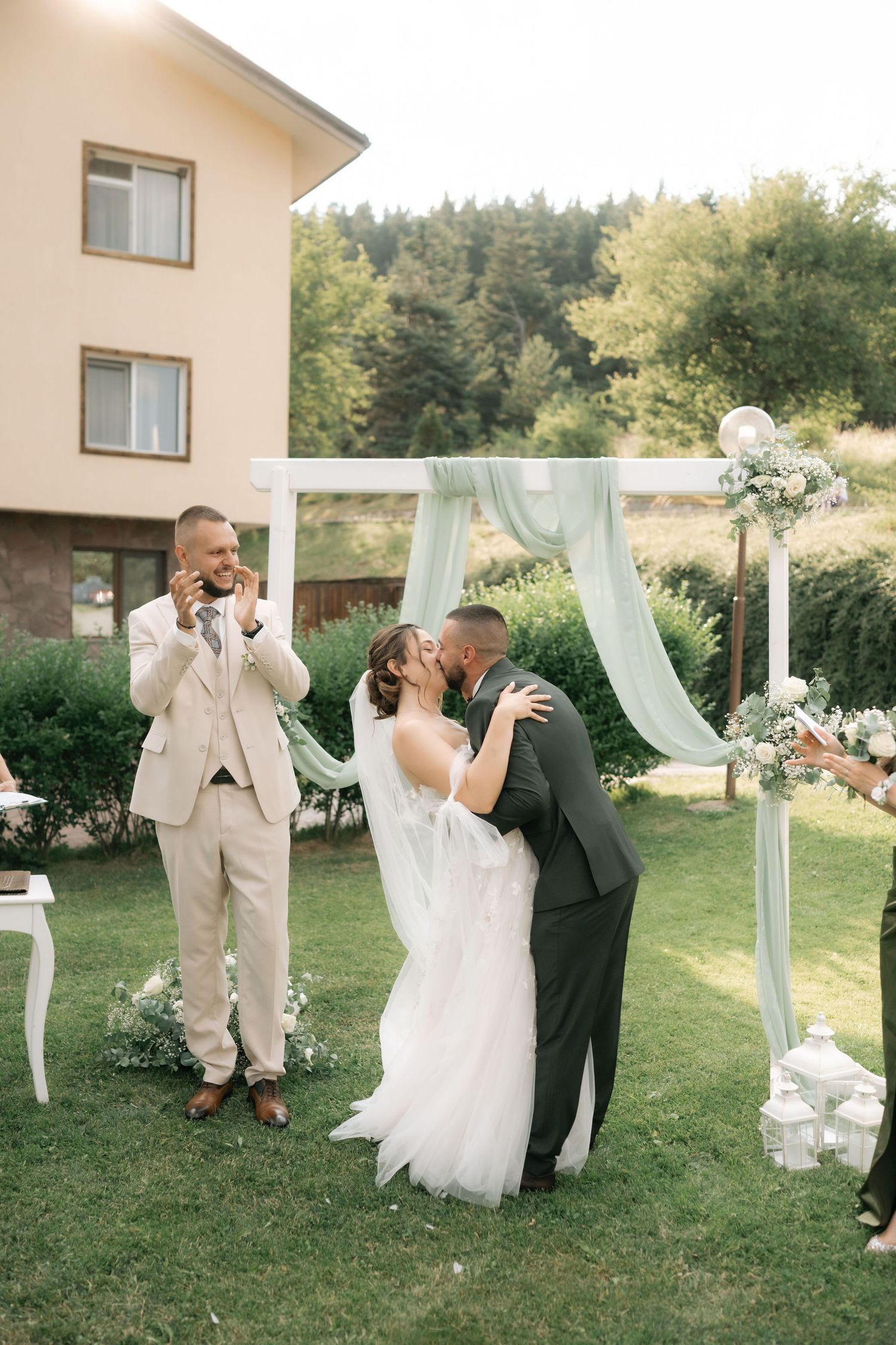 A couple shares their first kiss at an outdoor wedding ceremony under a light blue draped arch on a sunny day.