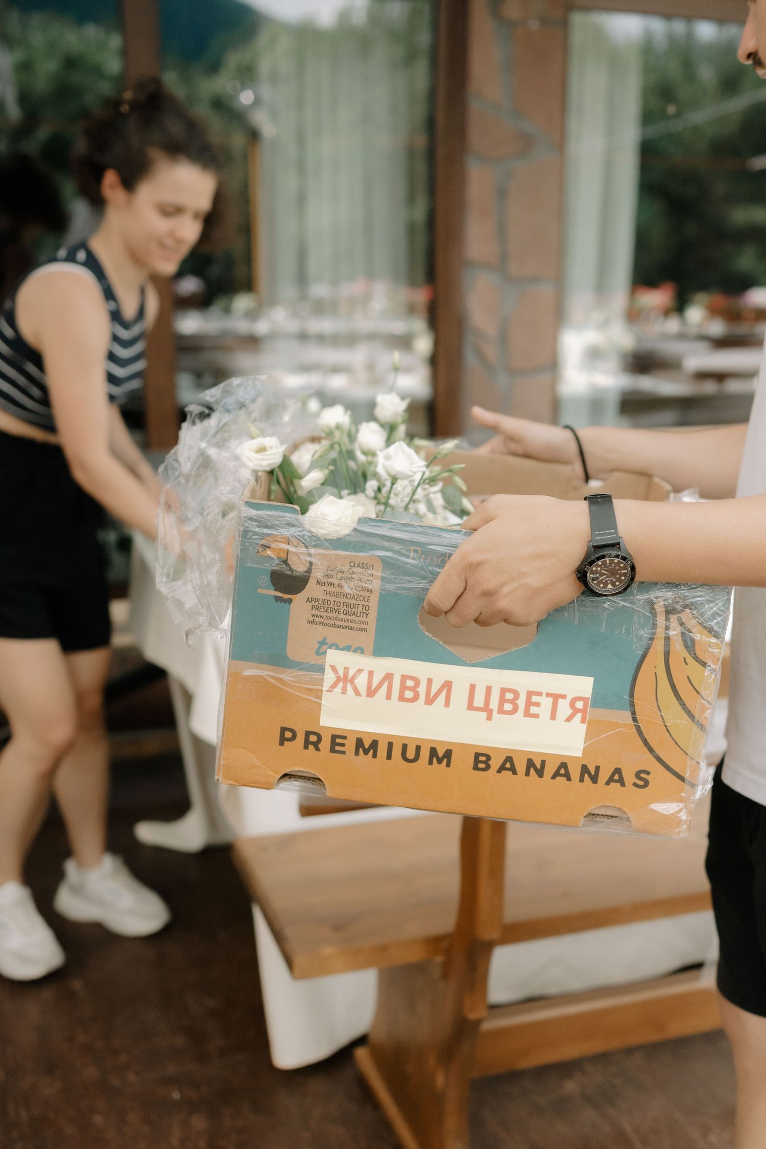 A cardboard box labeled 'Premium Bananas' with flowers on top being handled at what appears to be an event setup.