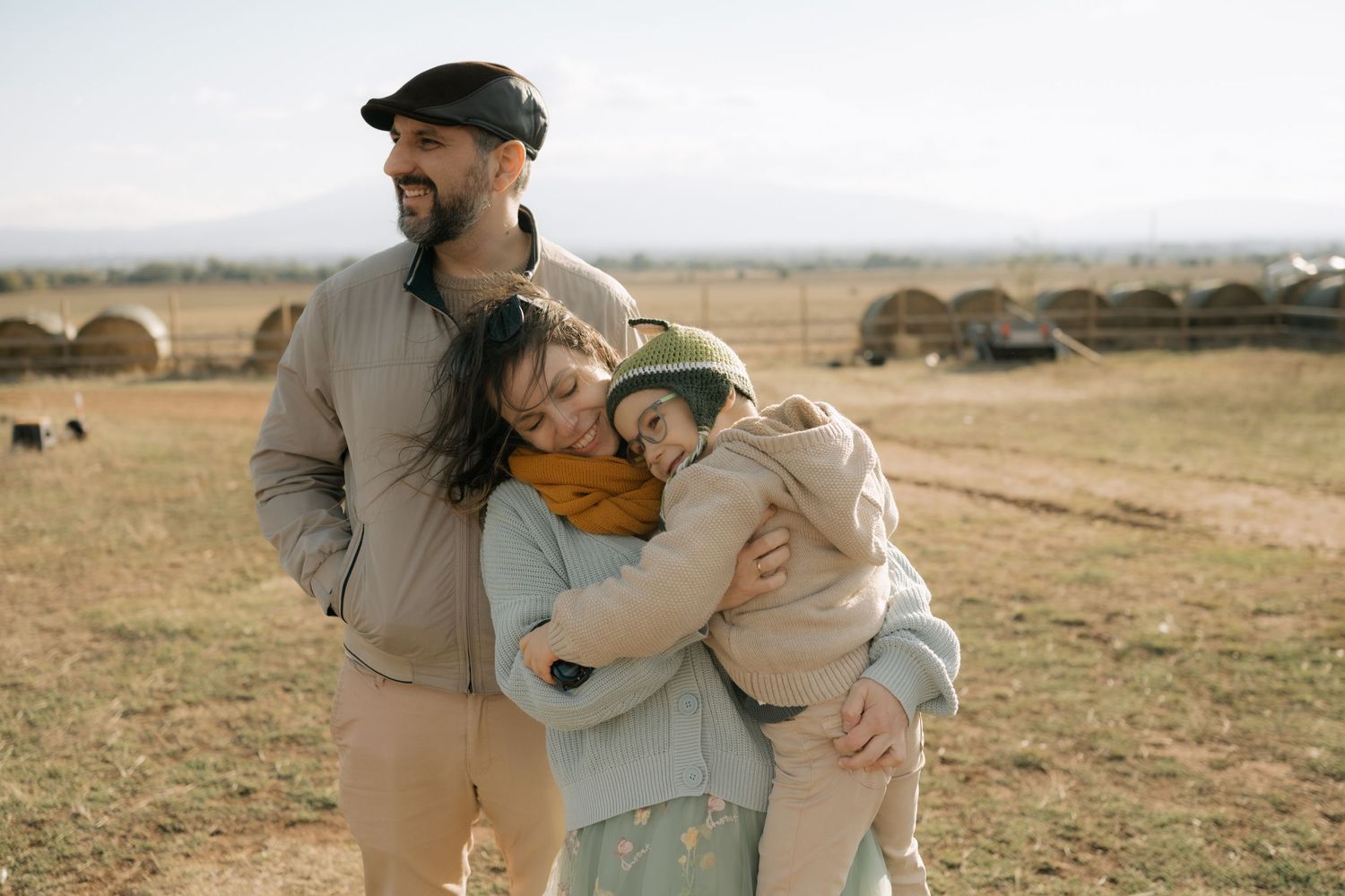 A family shares an embrace while standing in a field with round hay bales visible in the background.