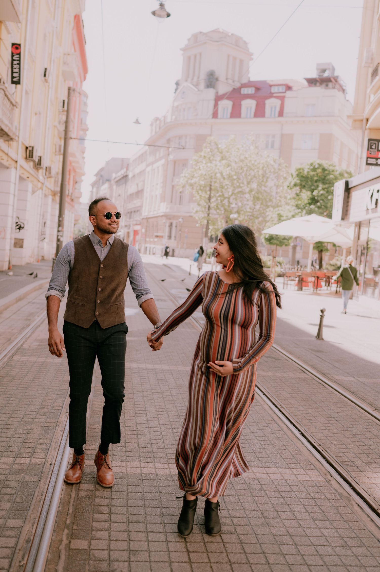 Couple holding hands walking on city street, woman in striped maxi dress, man in vest and sunglasses, urban background.