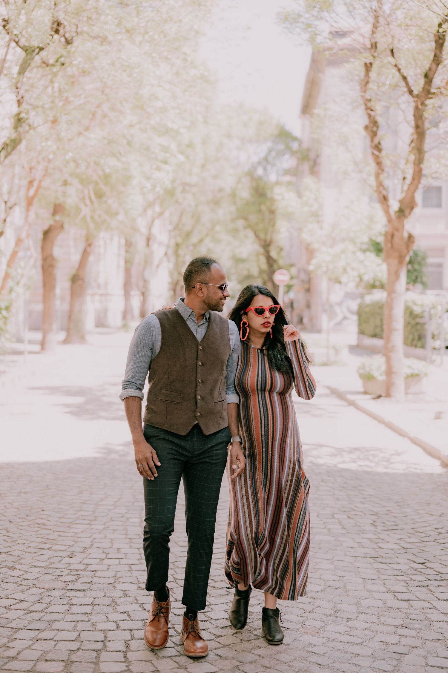A stylish couple walks on cobblestone path, man in vest and woman in striped maxi dress with sunglasses.