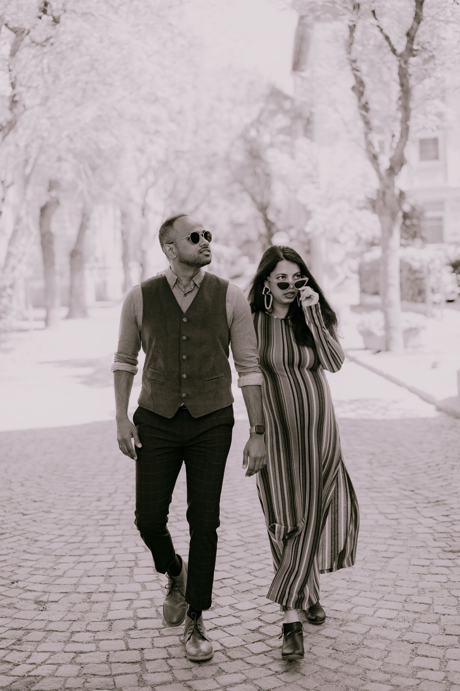 A stylish couple walks together on a cobblestone path, the man in a vest and the woman in a striped dress, both wearing sunglasses.