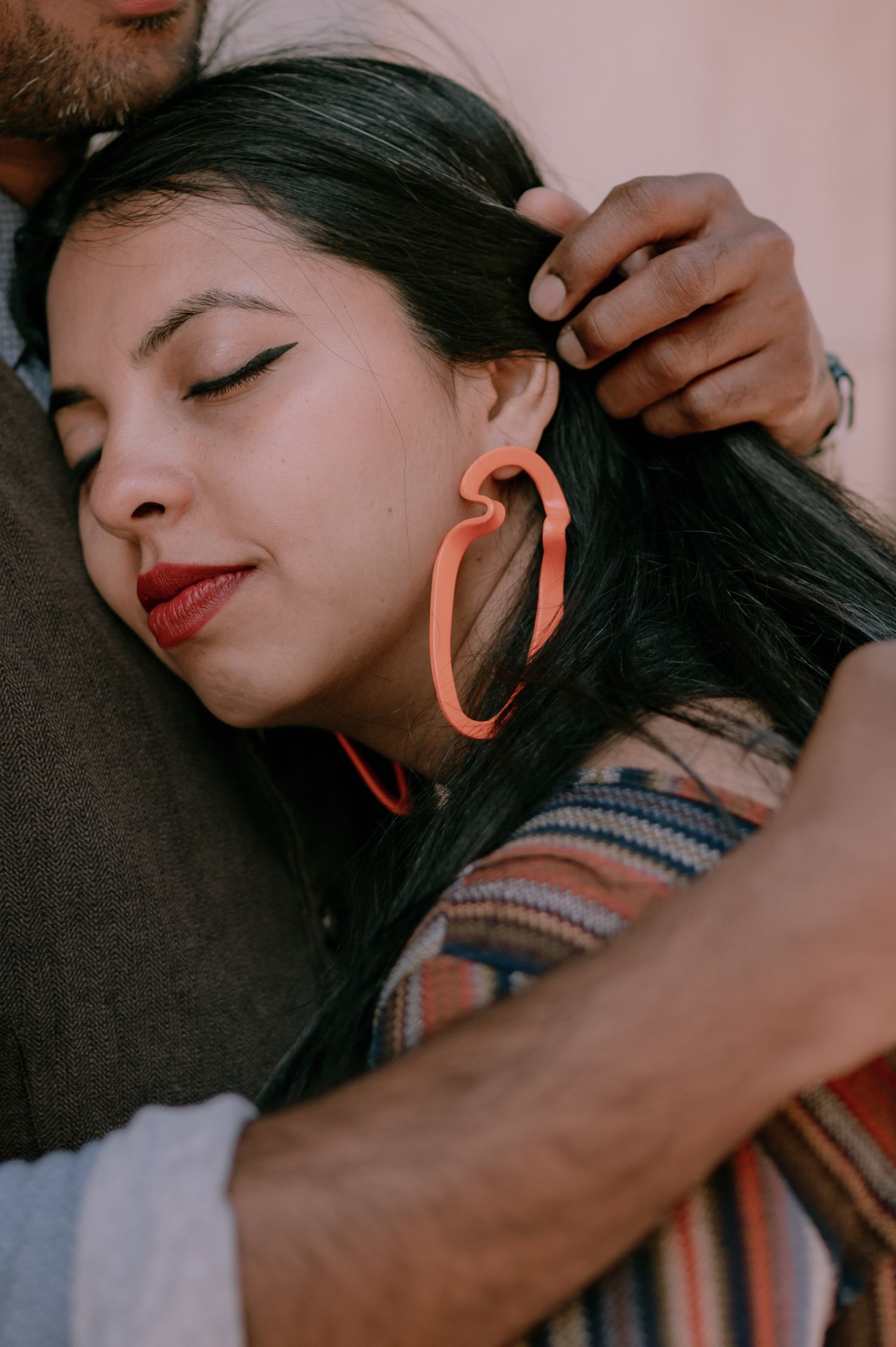 Woman with red lips and coral earring rests her head on a man's shoulder while he gently strokes her hair.