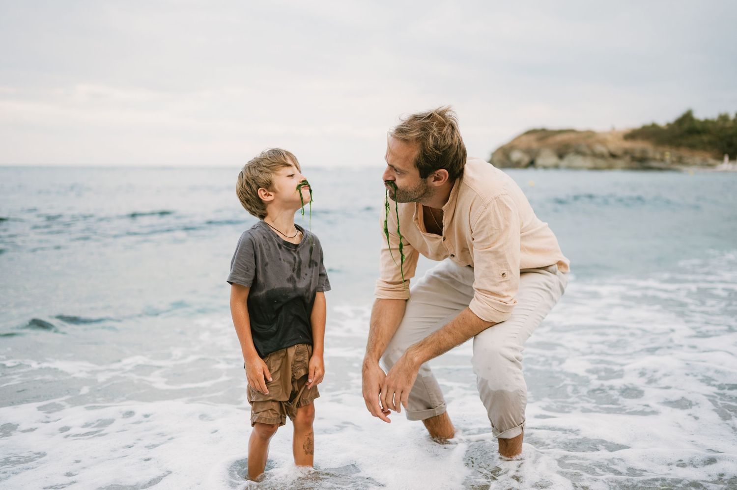 A father and young son laugh together while standing in ocean waves on a rocky beach.