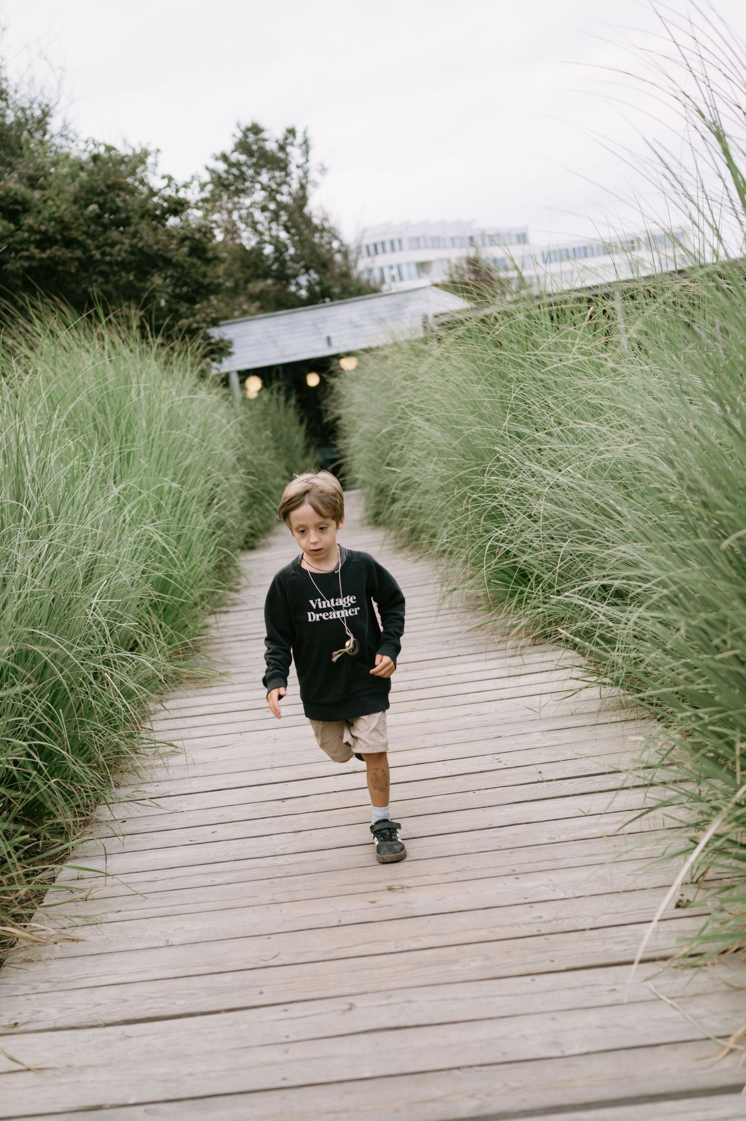 A young boy in a black sweatshirt runs along a wooden boardwalk path surrounded by tall green grass.