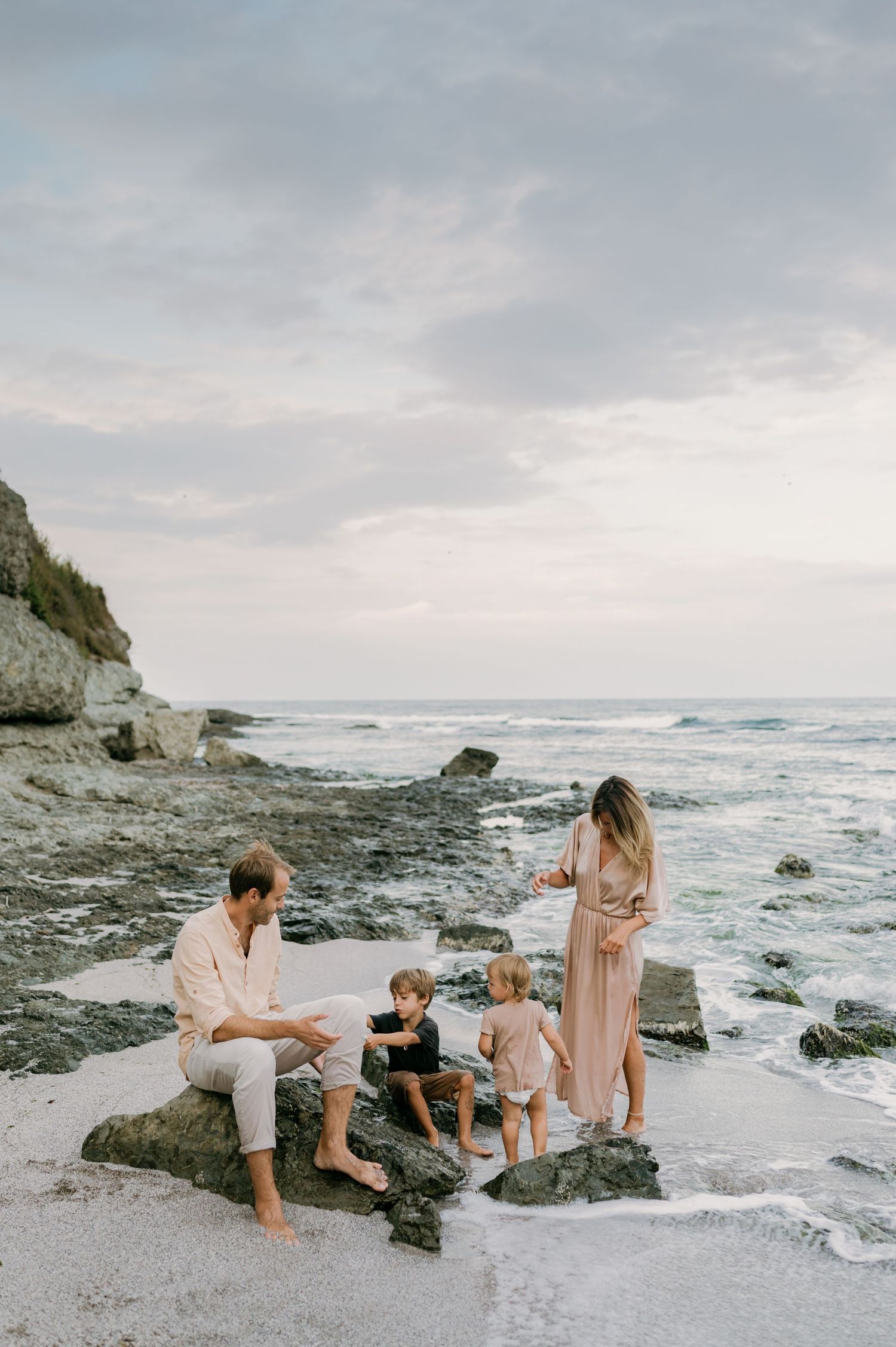 Family of four enjoying a rocky beach at sunset, with two young children playing near the ocean waves.