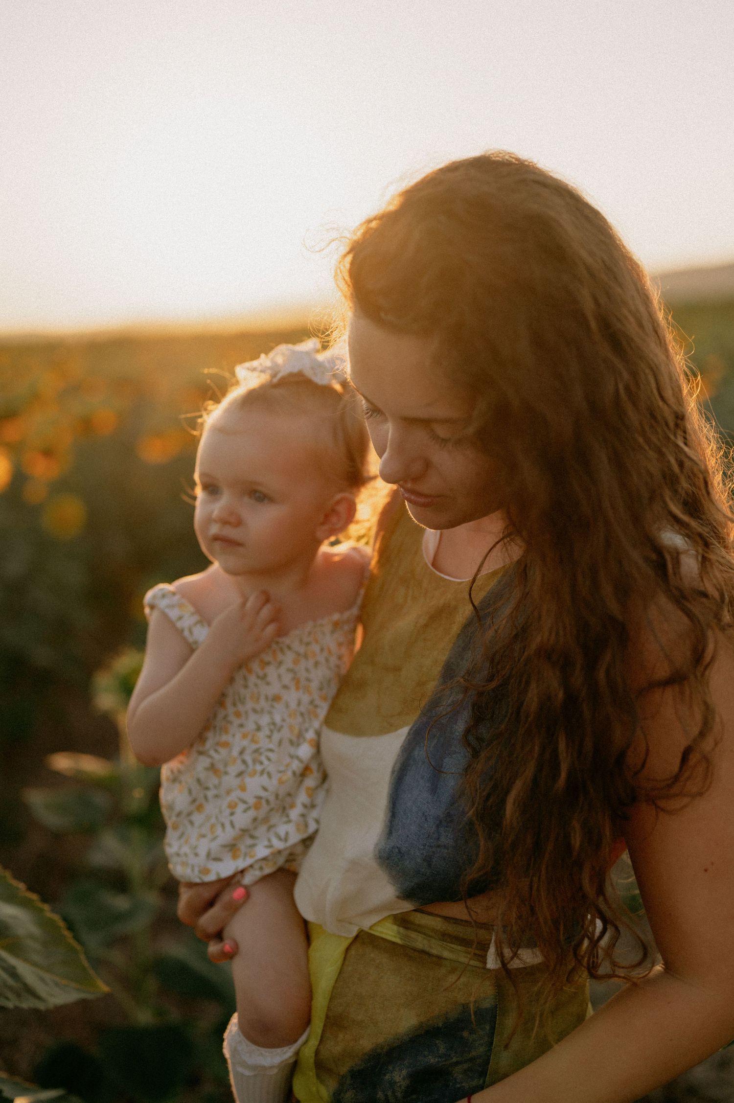 A mother with curly hair holds her baby girl in a sunflower field during golden hour sunset.
