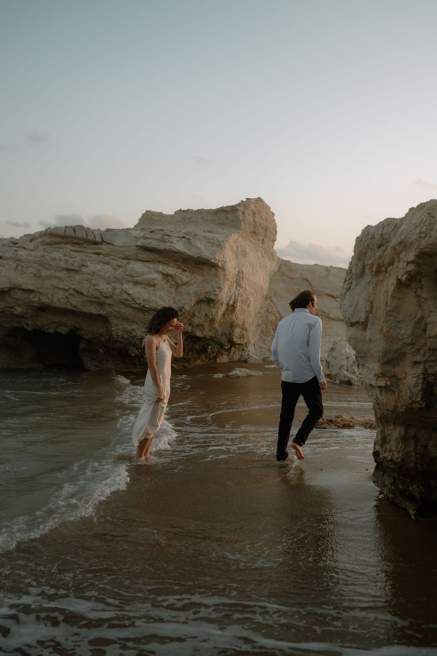 Couple walking barefoot on a sandy beach near white rock formations at dusk, woman in white dress, man in light shirt.