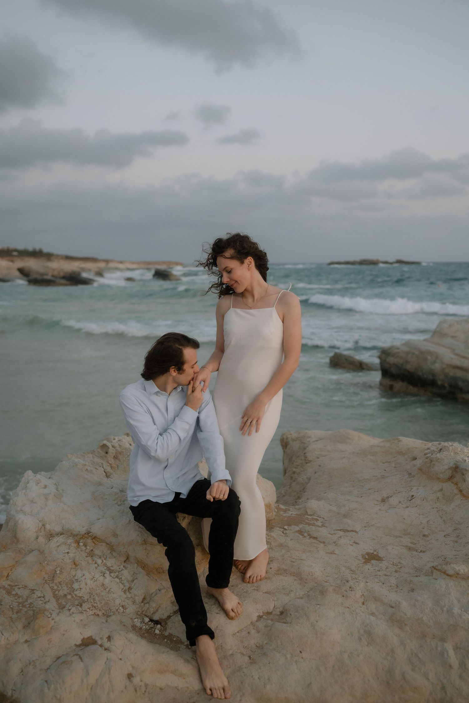 Couple sharing a romantic moment on coastal rocks, man seated holding woman's hand, ocean waves in background.