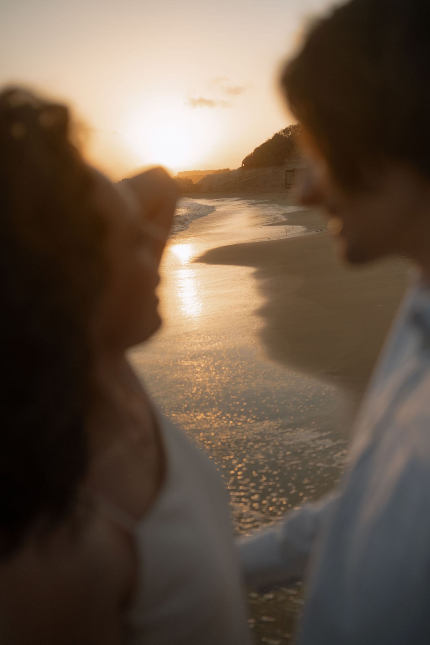 Couple sharing a romantic moment on a beach at golden sunset with ocean waves reflecting warm light.