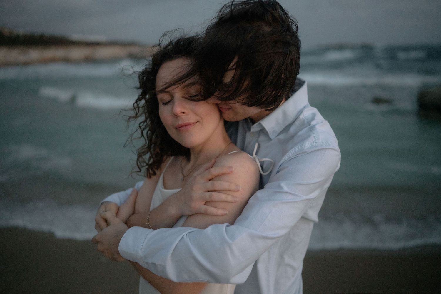 Couple embracing tenderly on a beach at dusk, waves crashing behind them in a romantic seaside moment.