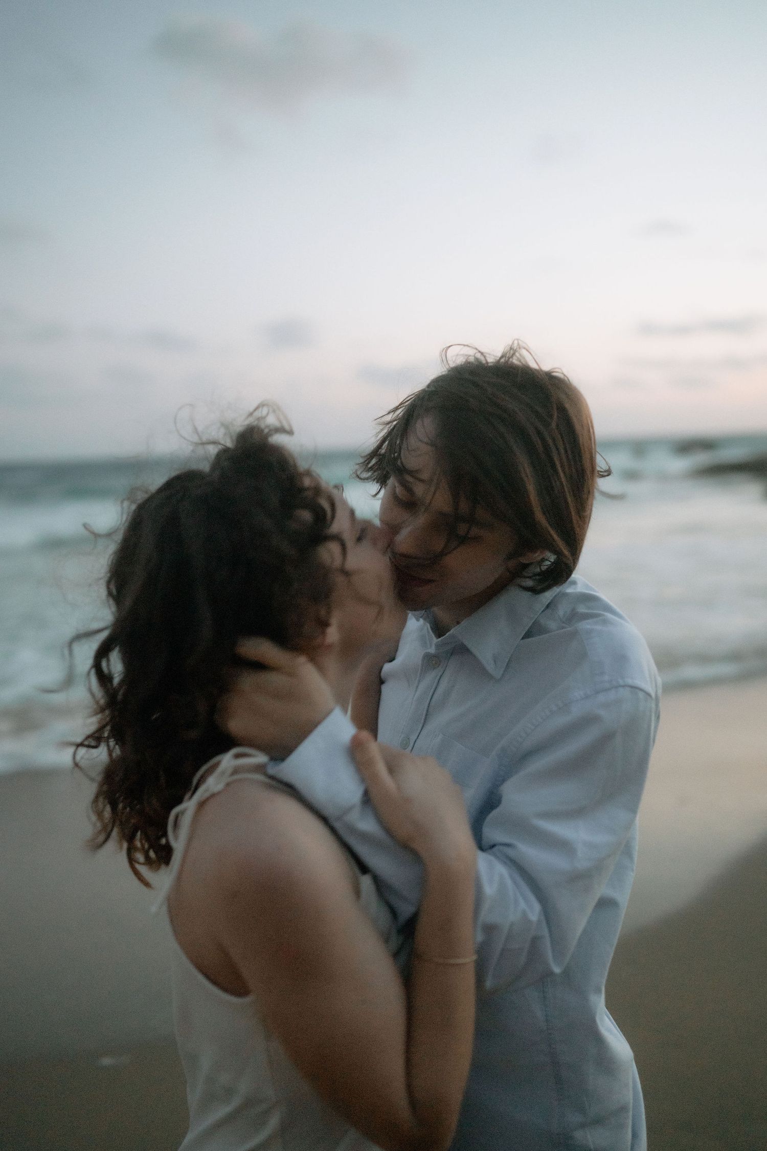 Romantic couple sharing an intimate moment on a beach at dusk, embracing with ocean waves in the background.