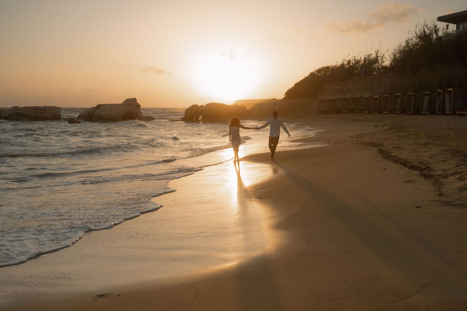 Couple walking hand in hand on a sandy beach at golden sunset with waves and rocky cliffs in the background.