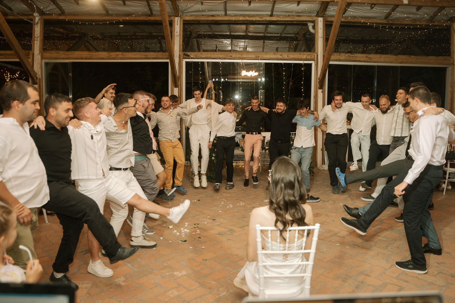 Wedding reception guests line dancing in a rustic barn venue with string lights overhead and wooden beams visible.