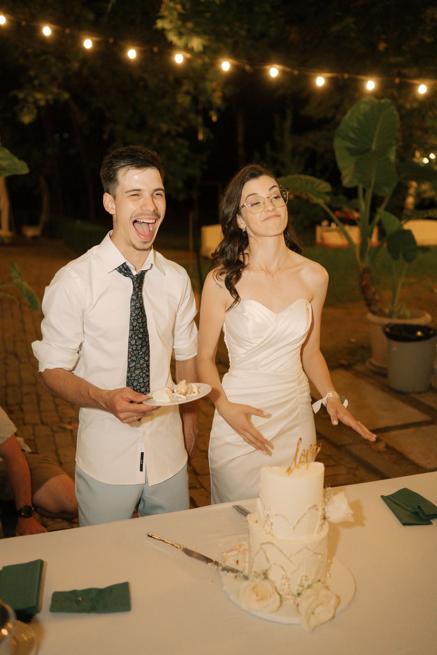 A couple laughs and shares a joyful moment while cutting their wedding cake together at an outdoor evening reception.