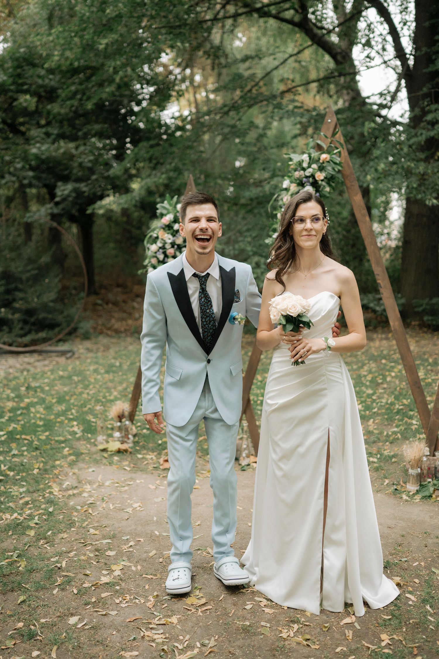 A bride and groom pose together in a wooded outdoor setting with fallen autumn leaves on the ground.