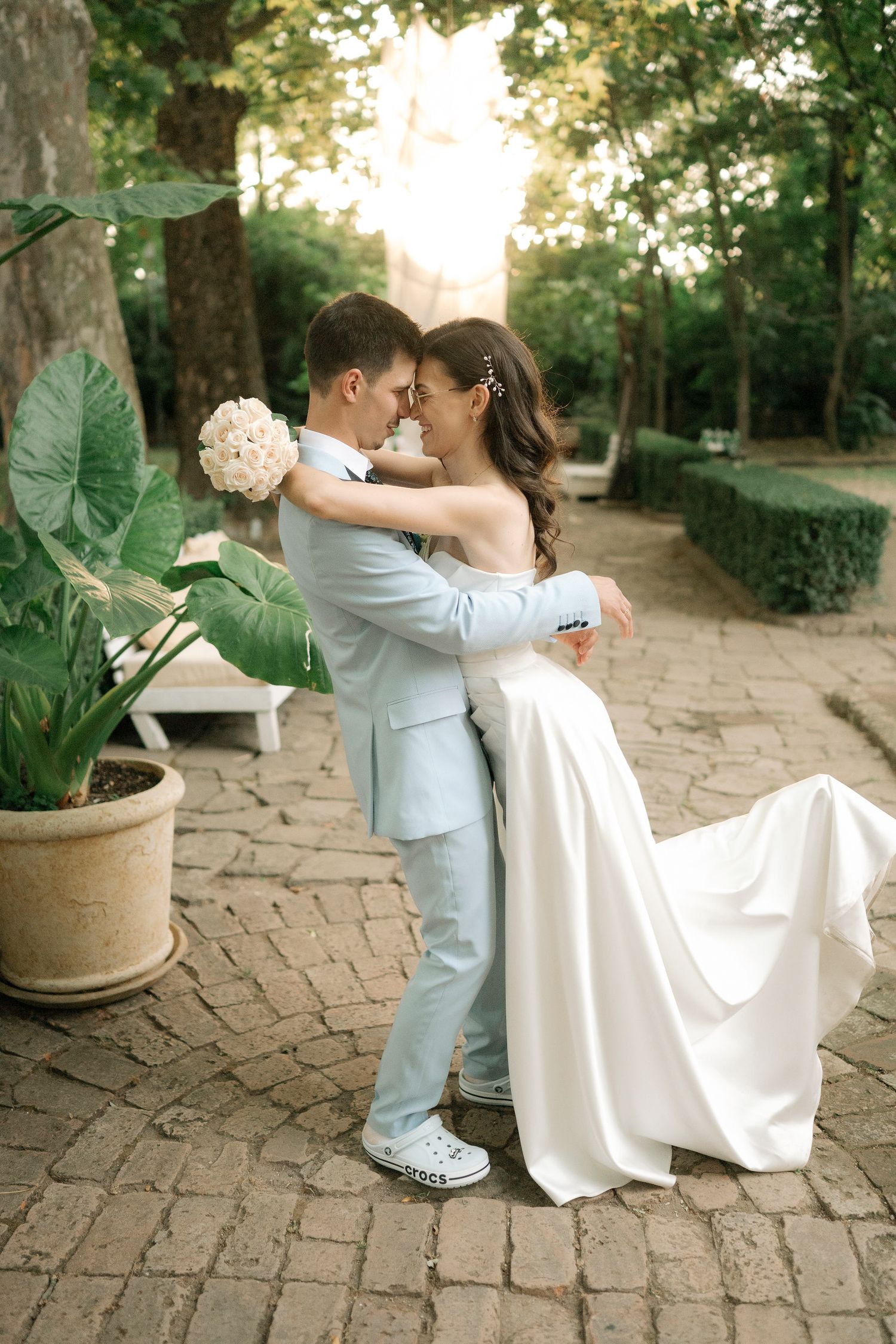 Romantic wedding couple embraces on brick pathway in garden setting with white bouquet and elegant attire at sunset.