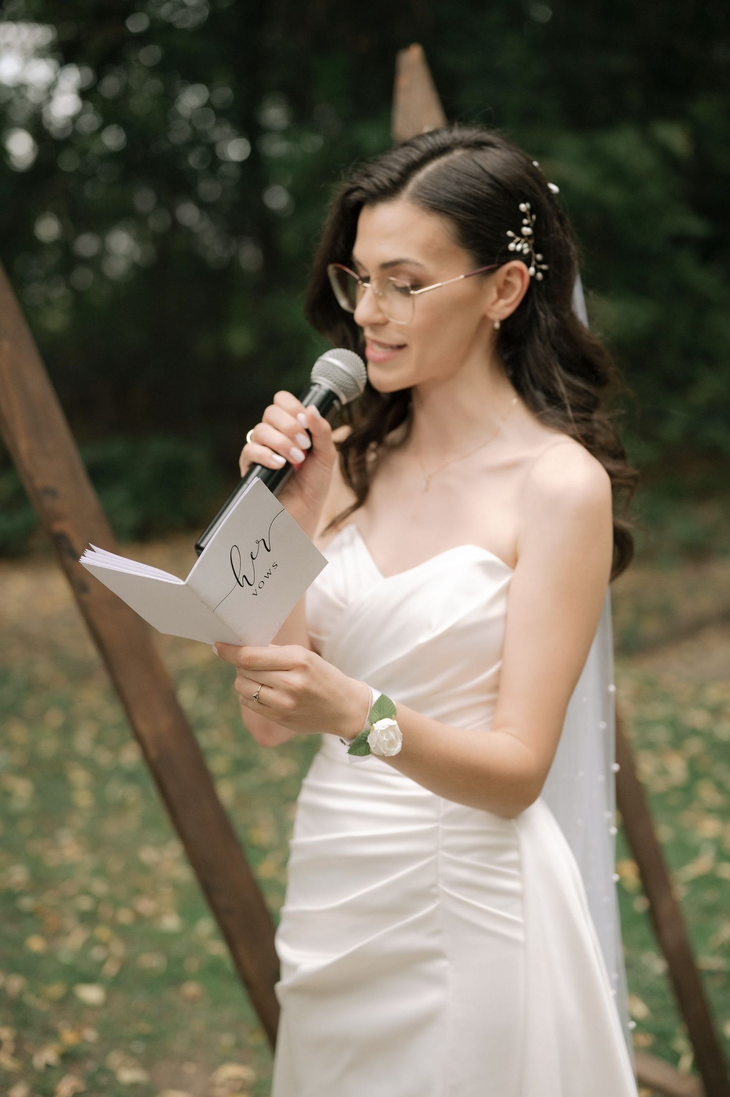 A bride in a white wedding dress reads from a paper during an outdoor ceremony in a wooded setting.