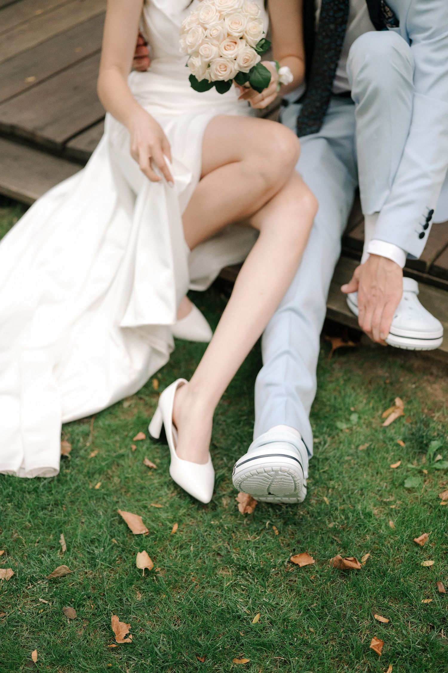A bride in white heels and groom in casual shoes sit on wooden steps with autumn leaves scattered on grass.