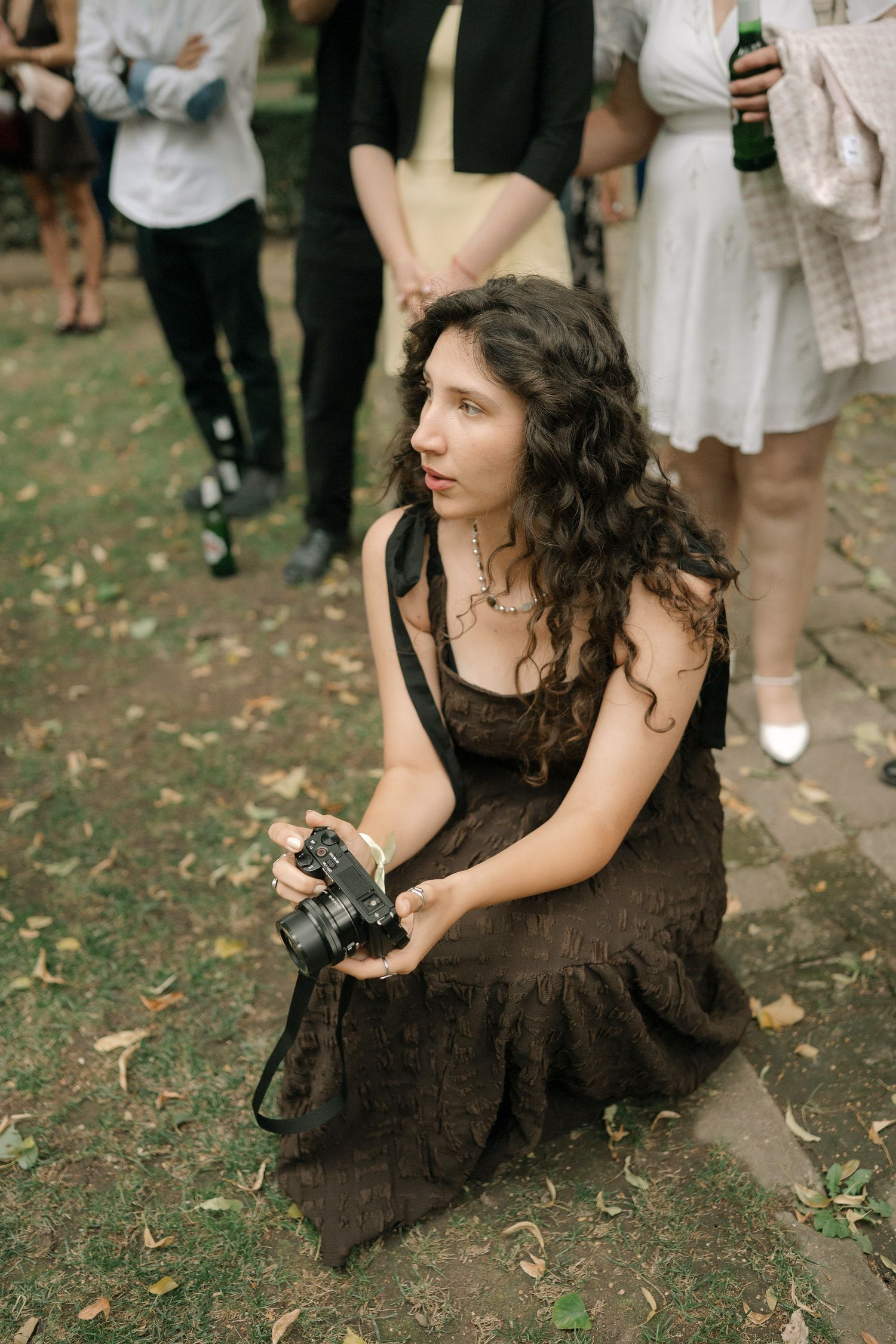 A photographer in a dark dress sits on grass while taking pictures at an outdoor event.