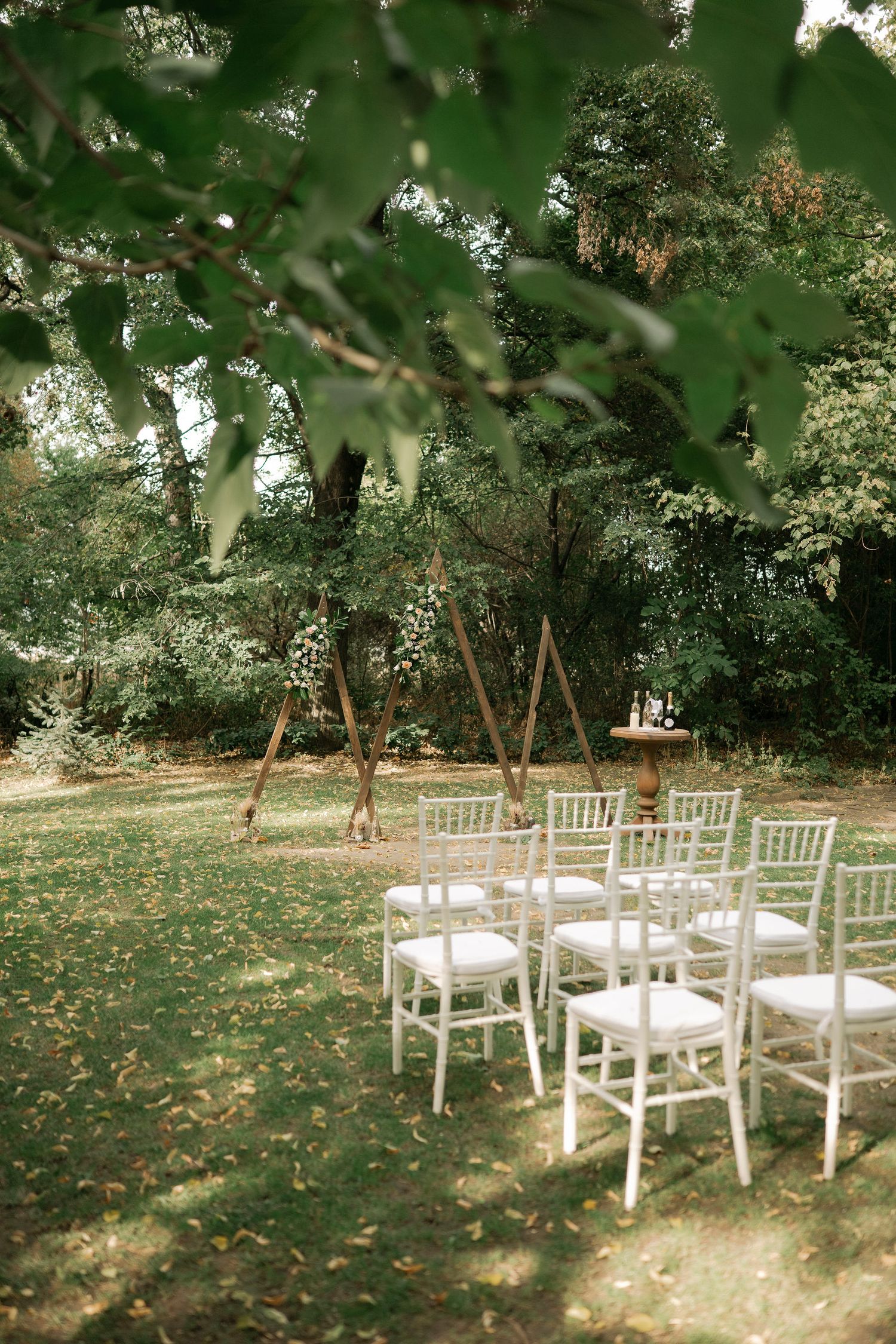 White wooden chairs arranged in rows on a grass lawn beneath shady trees for an outdoor garden ceremony.