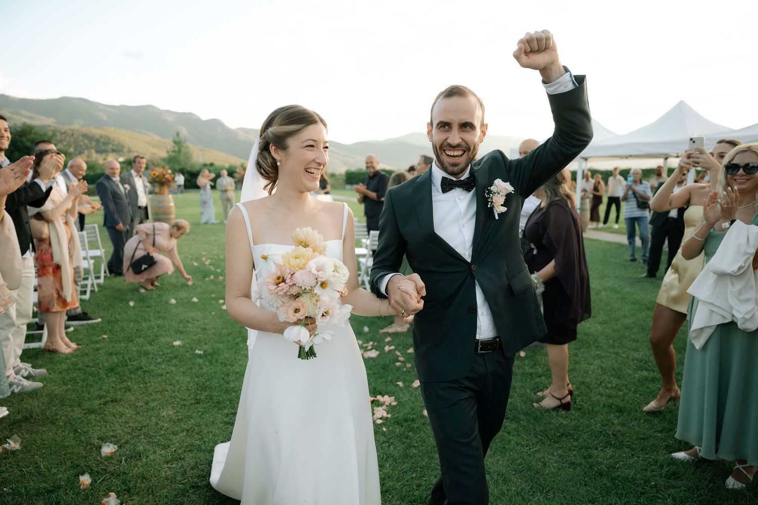 Wedding guests celebrate joyfully on a grassy hillside during an outdoor reception at sunset.