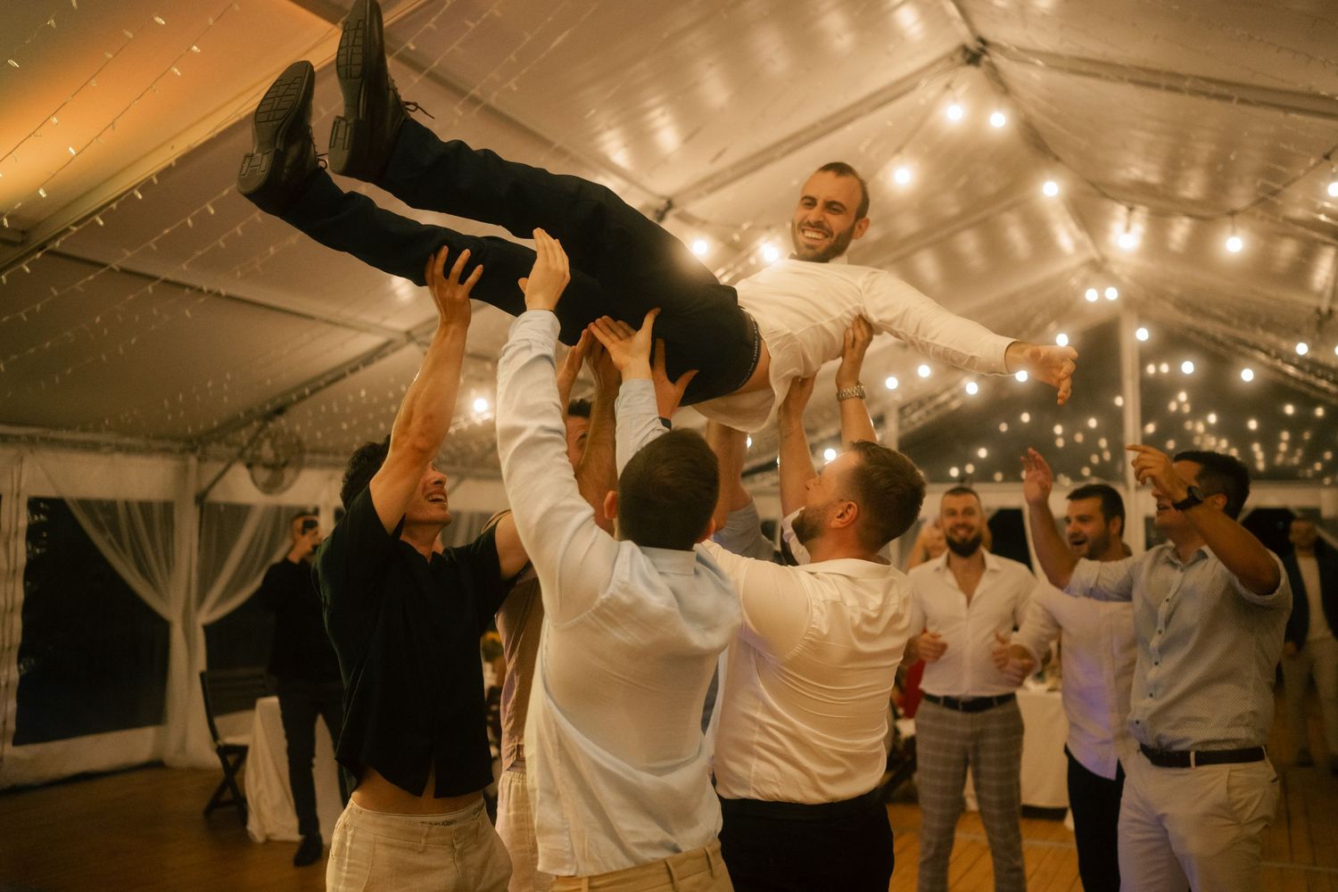 Wedding guests dance and celebrate under a white tent with string lights during an evening reception.
